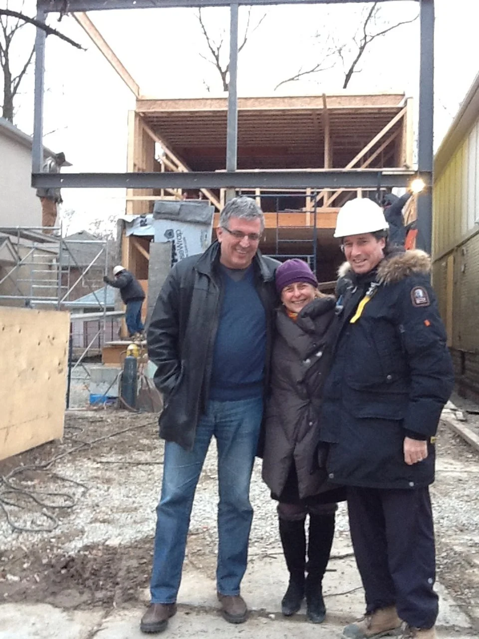 Three people smiling at a construction site, with a partially built wooden house in the background. Two men and one woman are in the foreground, wearing warm clothing and hard hats, indicating they are at a building or renovation project.