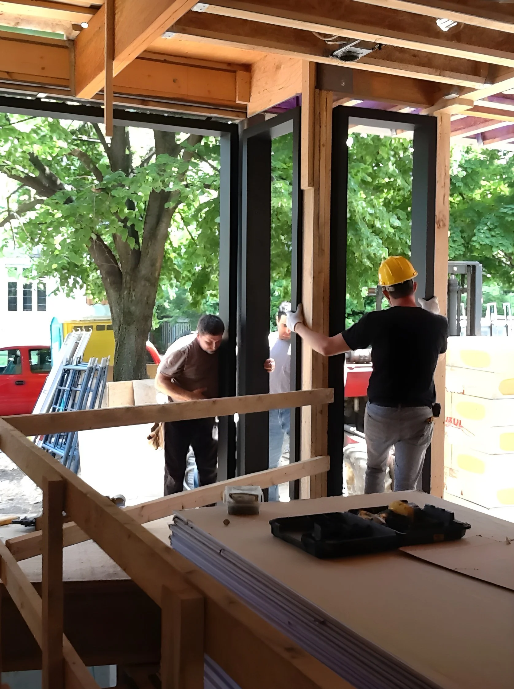 Construction workers installing large window frames in a building under construction with wooden framing and a view of green trees outside.
