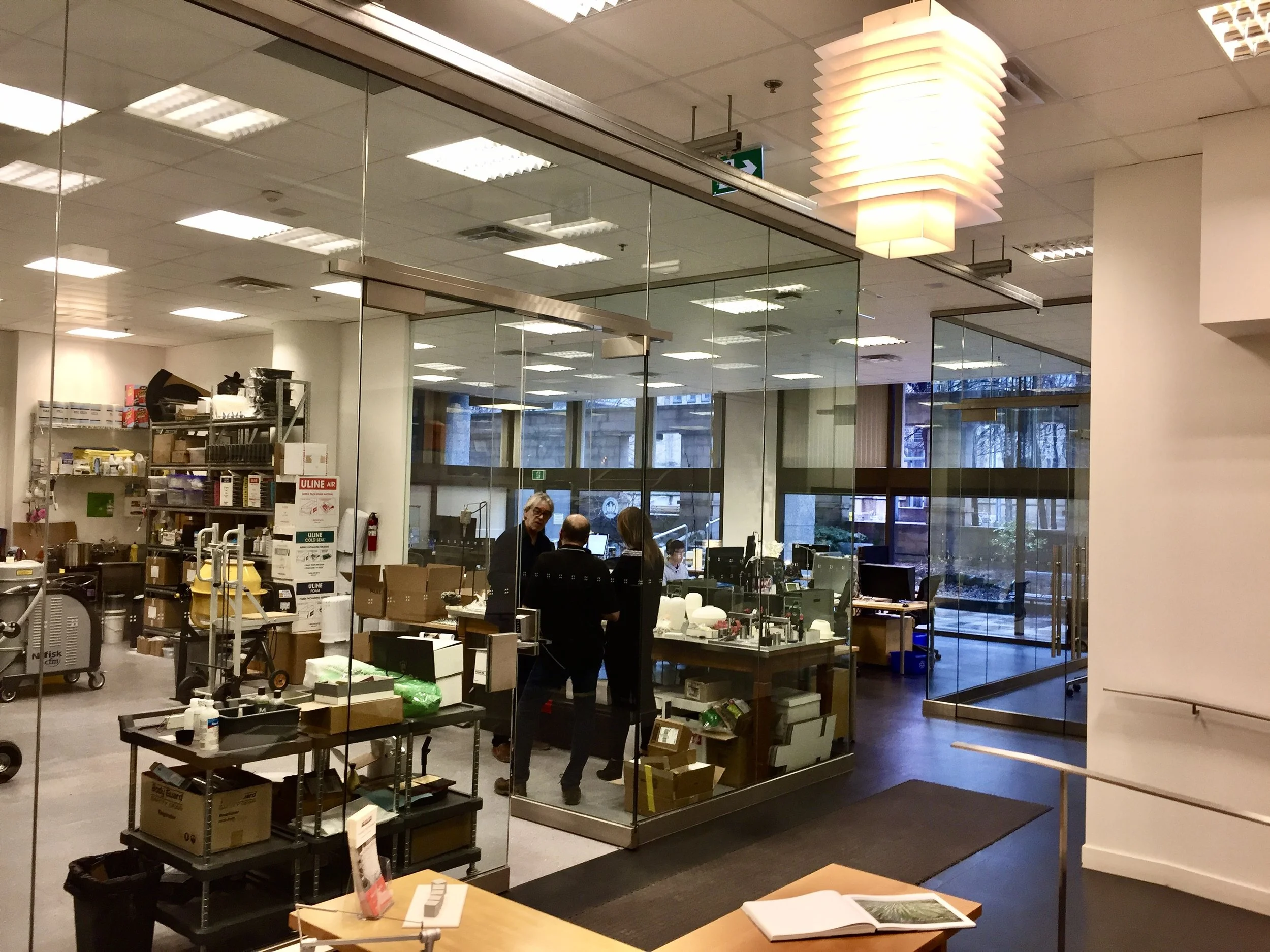 Interior of a 3D printing lab with glass walls, showing three people in conversation, and a view of an outdoor area through large windows.