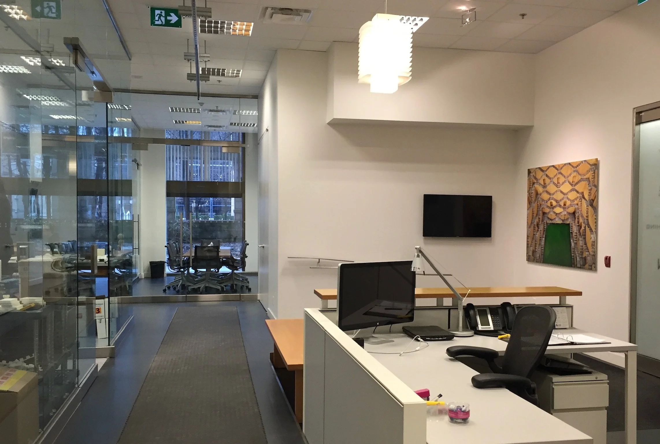 Office reception area with a desk and a large photograph on the wall. Behind the desk are glass doors leading to a conference room with chairs. There is a ceiling light fixture and exit sign.