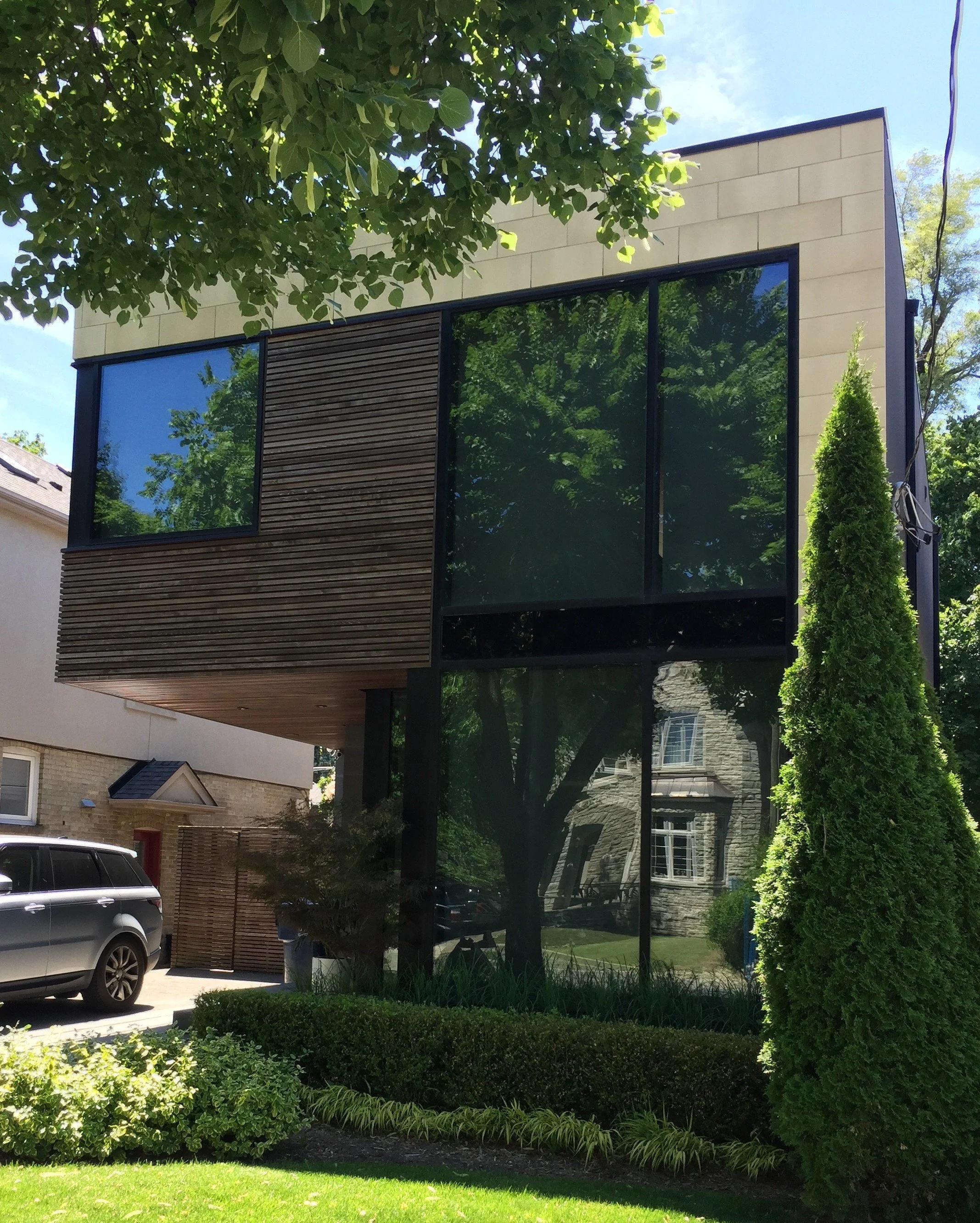 Modern two-story house with large glass windows, wooden paneling on part of the exterior, and surrounded by green trees and shrubs.