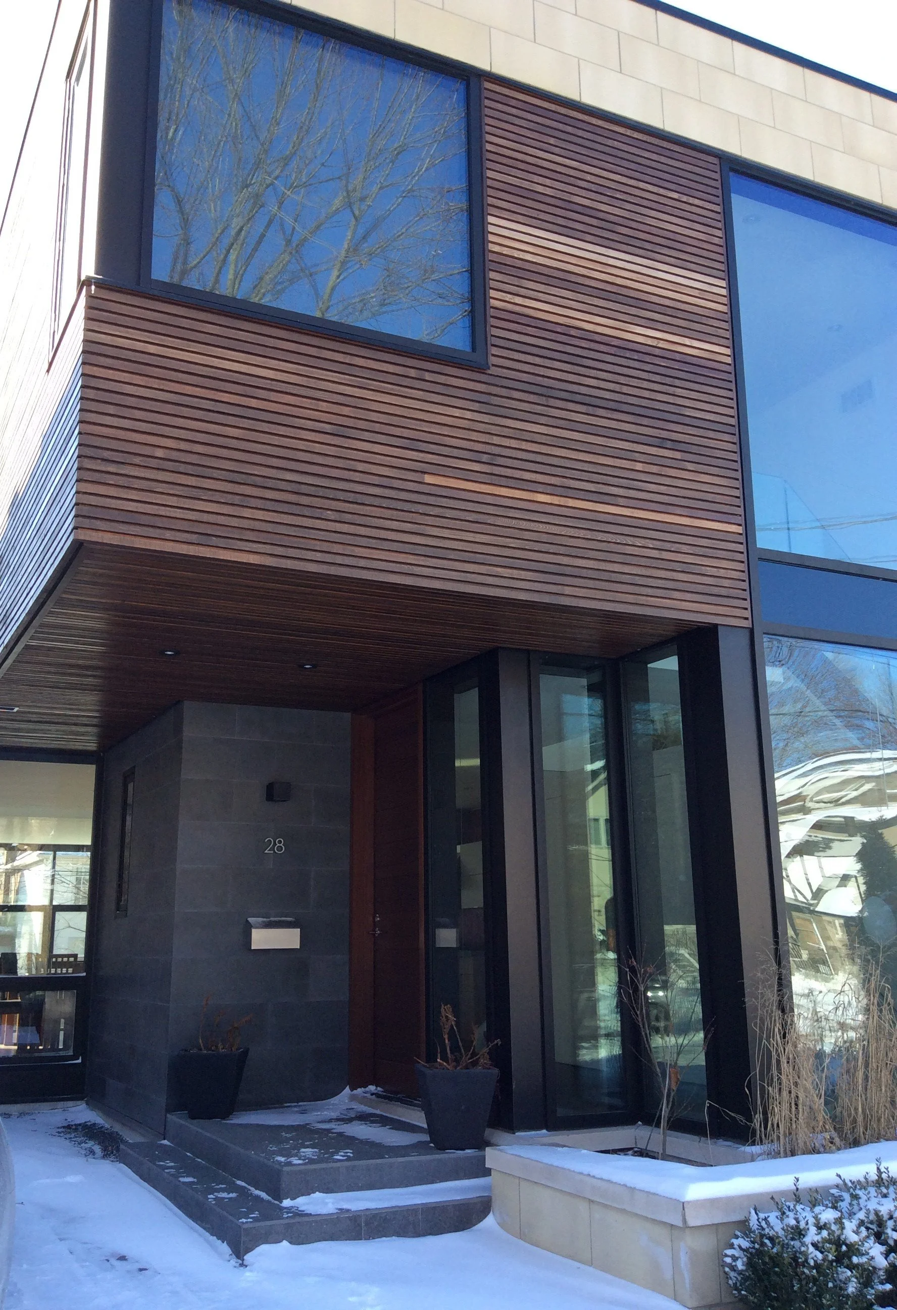 Modern house with dark gray stone exterior, wooden siding, large glass windows, and a small snow-covered front porch with planters.
