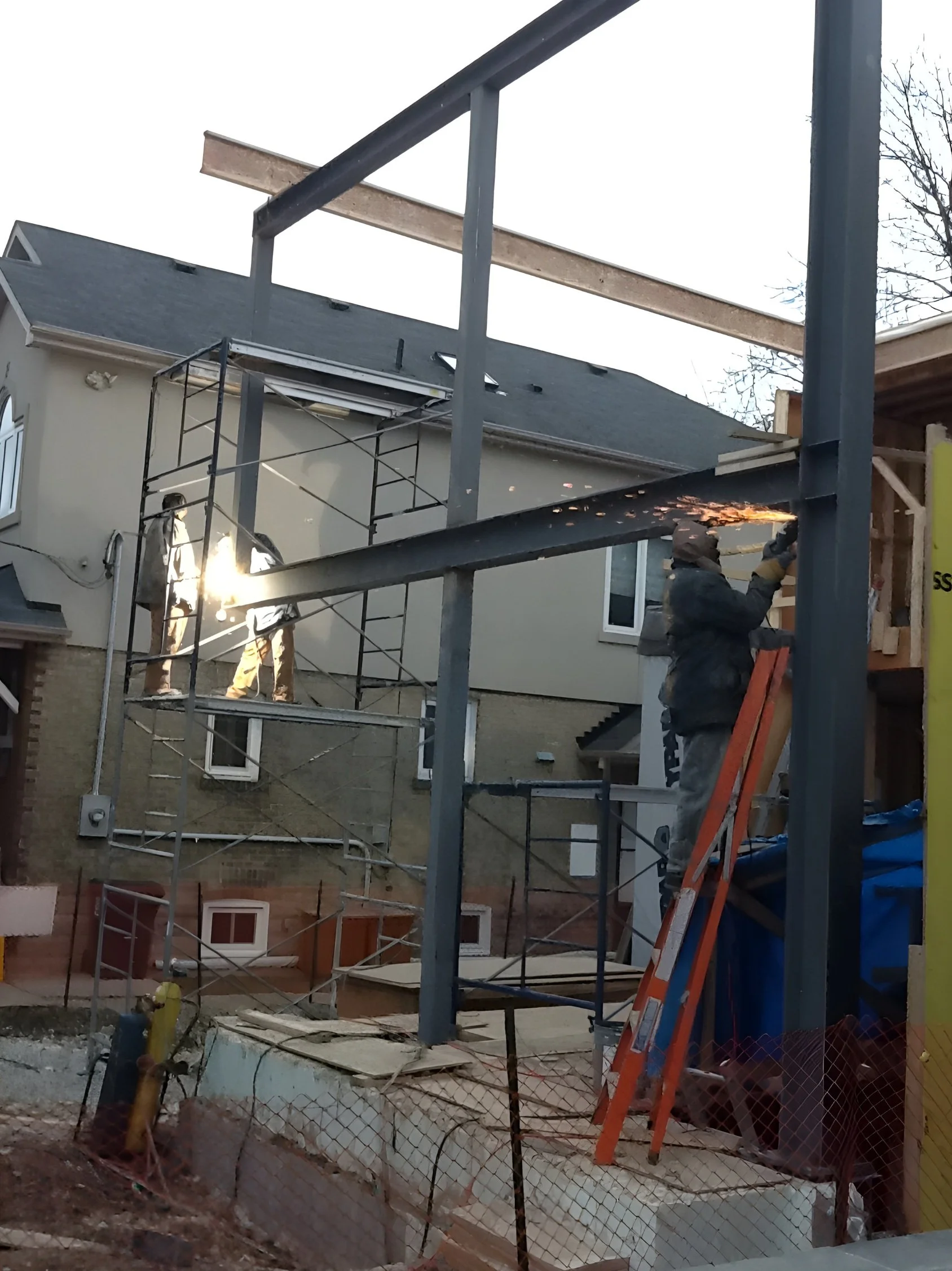 Construction workers installing horizontal metal beams on a building frame, with one worker welding sparks visible, in a residential area with scaffolding and a house in the background.