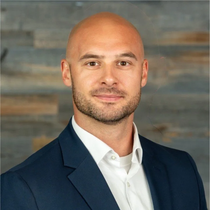 A portrait of a bald man with a beard wearing a navy blue suit jacket and white shirt, standing against a wooden wall background.