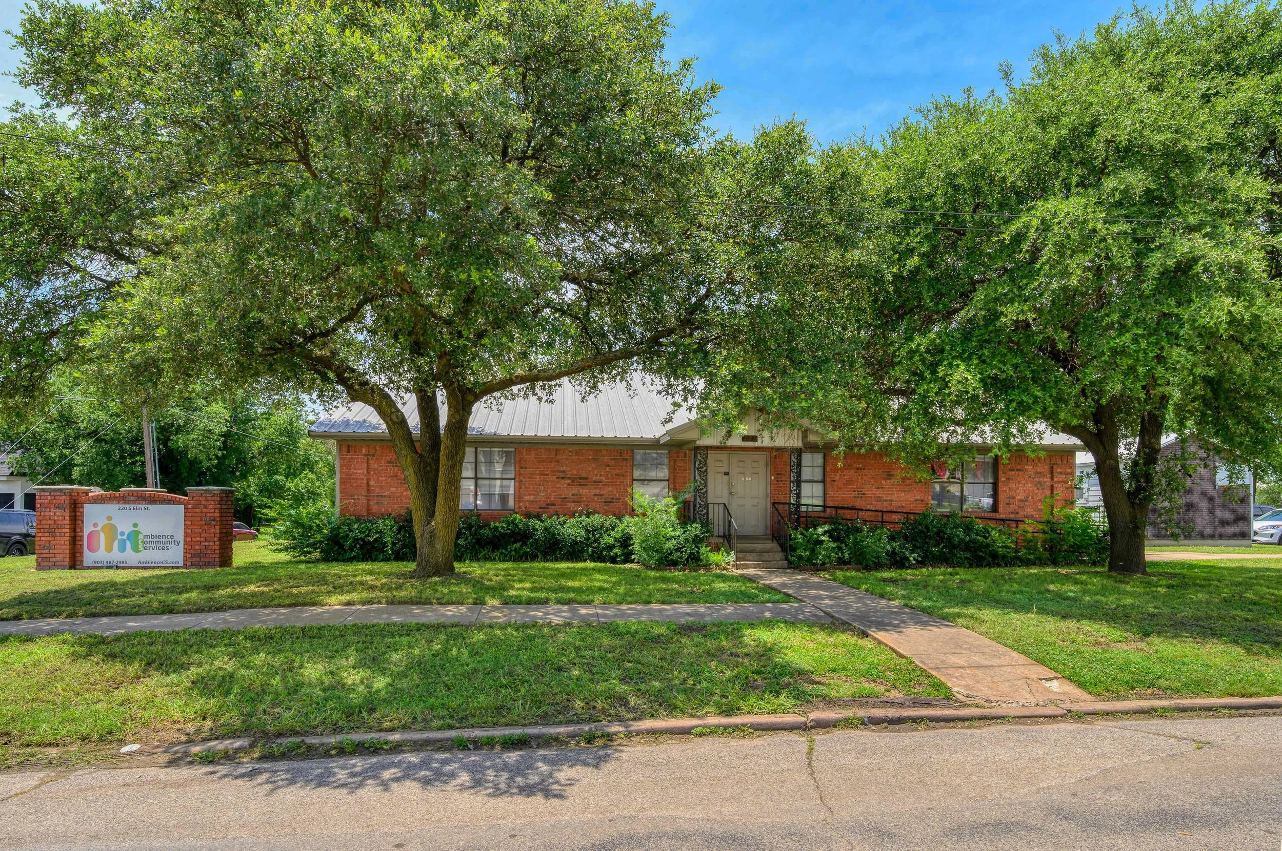 A brick building with a light gray roof, surrounded by green trees and grass, with a sidewalk in front. There is a sign on the left reading 'Ambiance Community.'