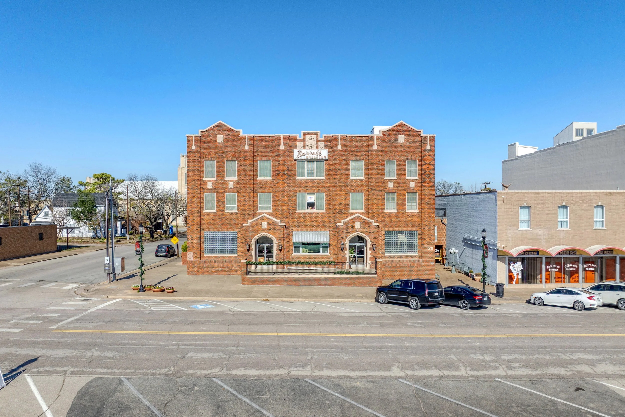 A three-story brick building with decorative brickwork, large windows, and a sign that reads 'Barnett Building'. The building is on a street with parked cars, planters, and a tree-lined sidewalk, with a clear blue sky overhead.