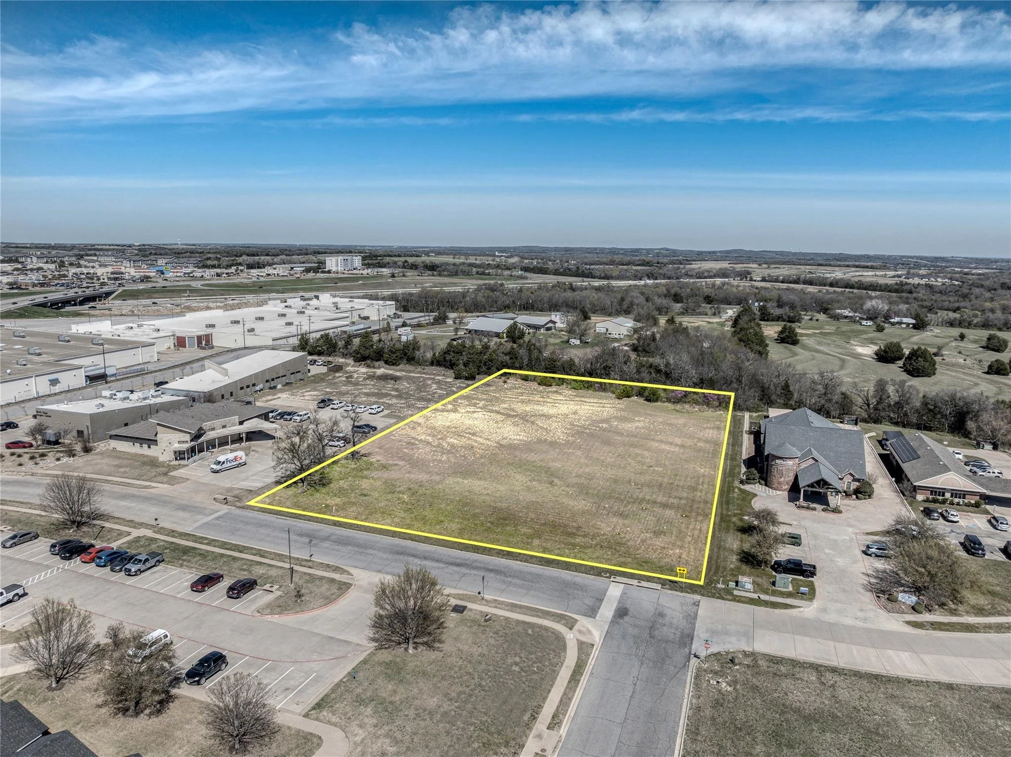 A vacant lot outlined in yellow in a suburban area, surrounded by parking lots, commercial buildings, and residential houses, with trees and open land in the background under a blue sky with wispy clouds.