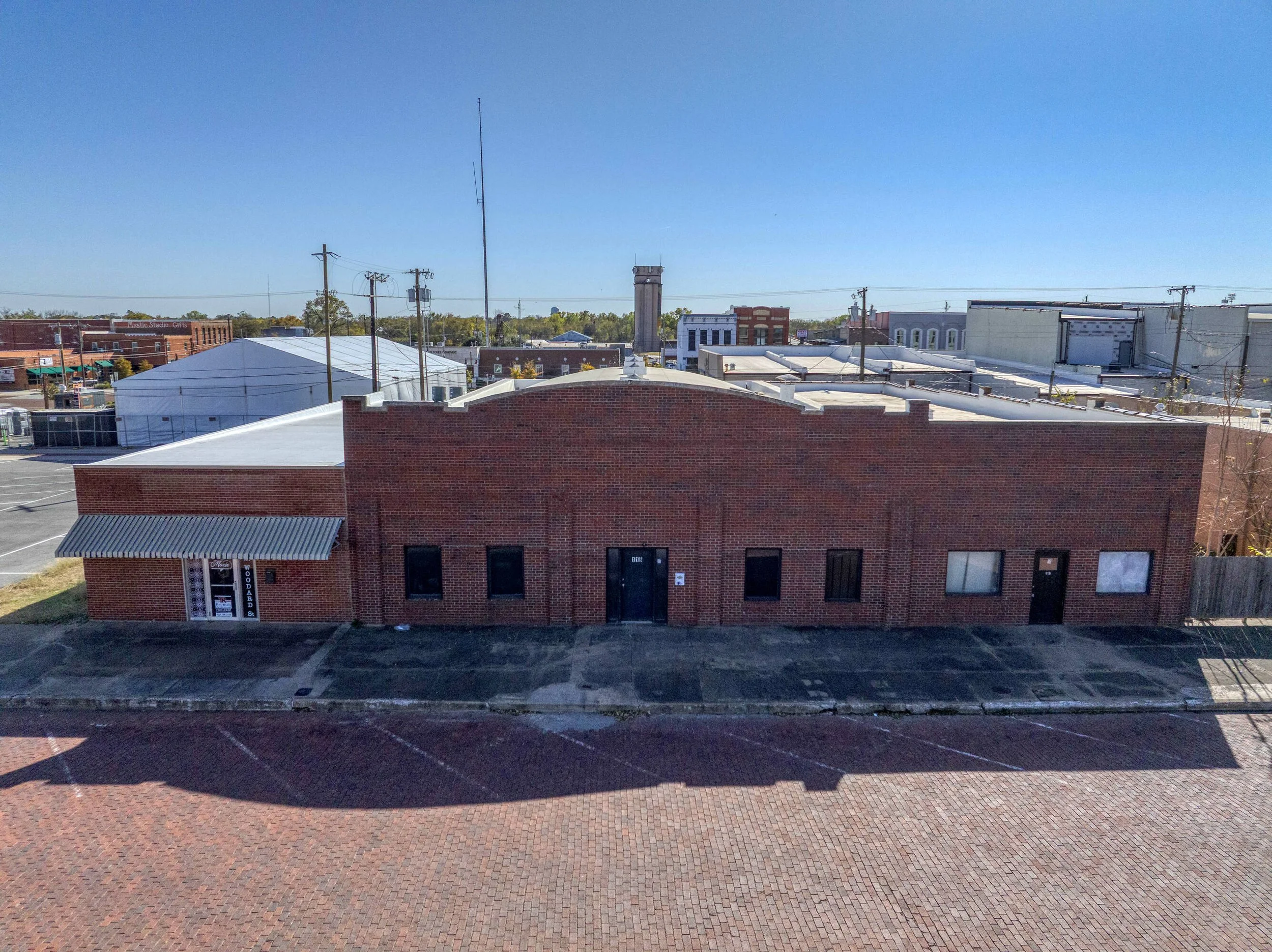 A brick commercial building with a metal awning and parking lot in front, located in a downtown area on a clear day.