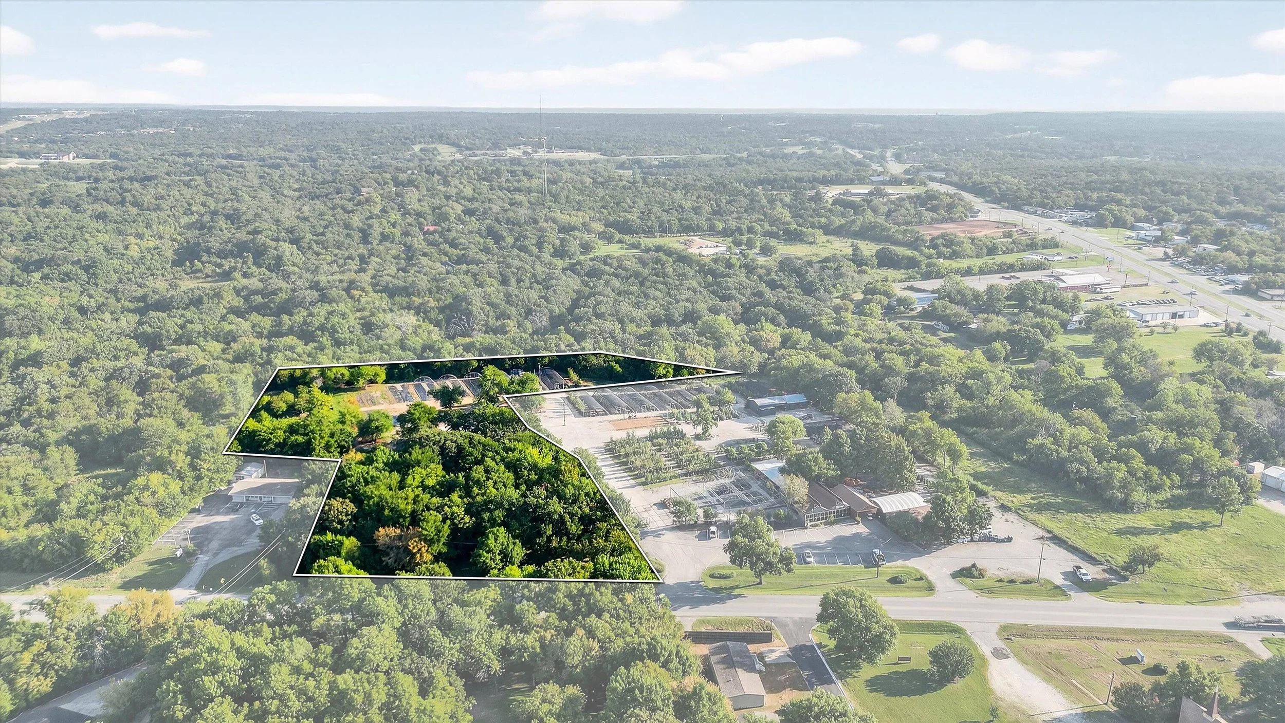 Aerial view of a large property outlined in white with dense green trees, parking lots, and surrounding daytime landscape.