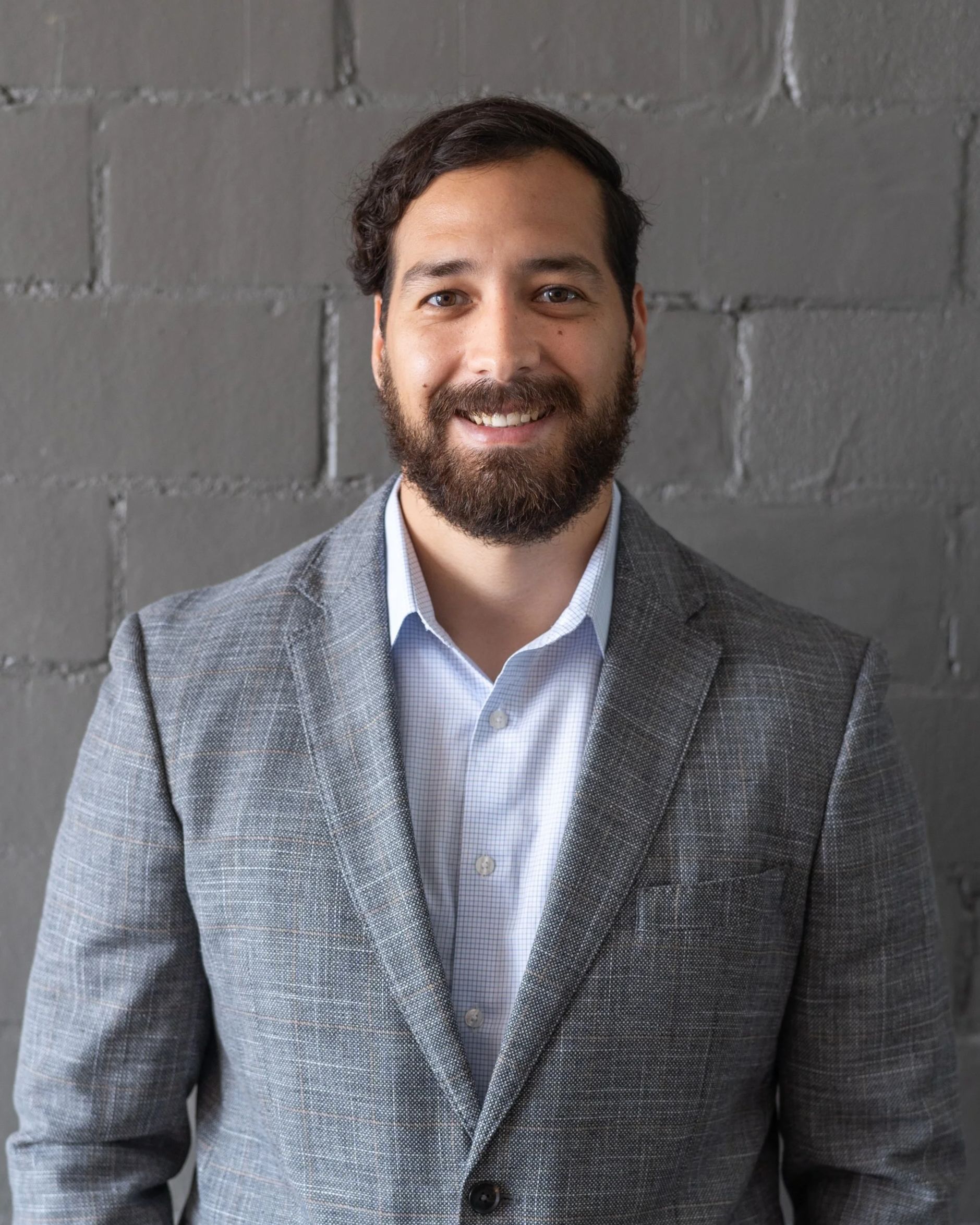 A man with a beard and dark hair smiling in front of a gray brick wall, wearing a gray plaid suit jacket and a white shirt.