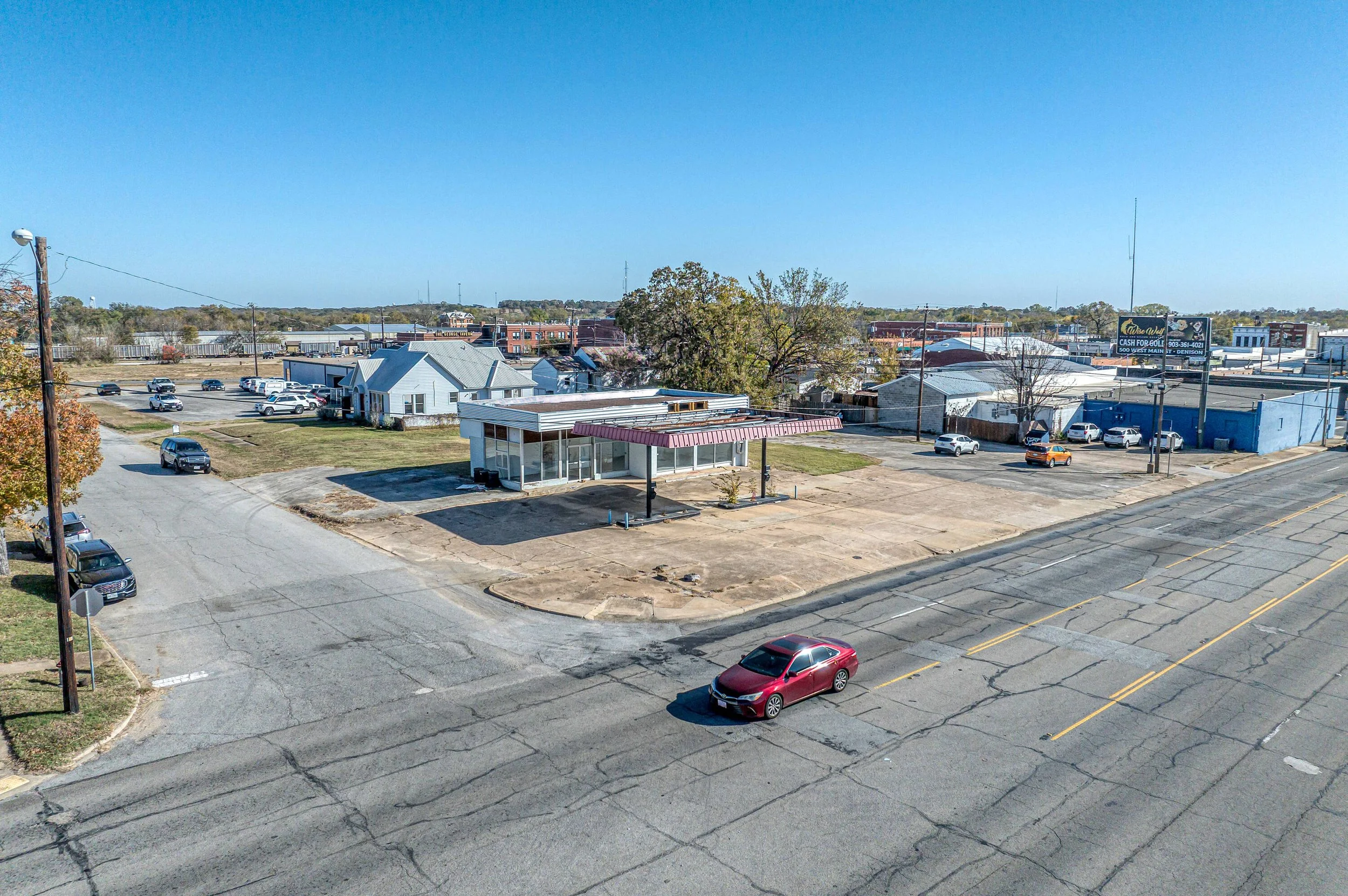 A deserted small town street scene with a closed fast food restaurant, a parking lot with few cars, and a few small buildings in the background under a clear blue sky.