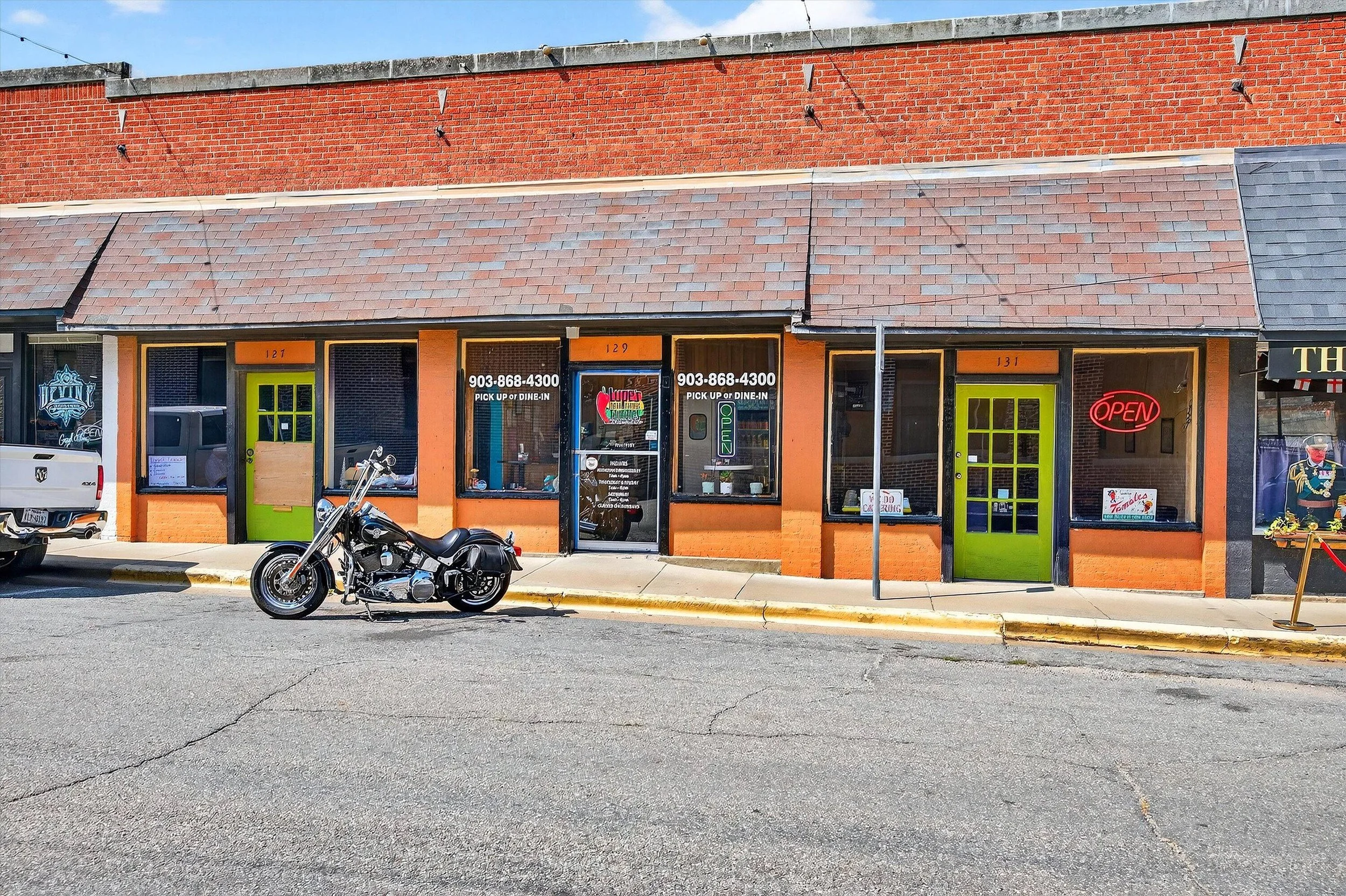 A small strip mall with three storefronts. The middle restaurant has large windows, a glass door, a neon 'OPEN' sign, and a sign with the phone number 903-868-4300. The storefronts on each side have lime green doors, and the one on the right has a red neon 'OPEN' sign. A black motorcycle is parked in front, along with part of a white pickup truck on the left. The building has brick walls with a sloped shingle roof, and the sidewalk is in front of the stores.