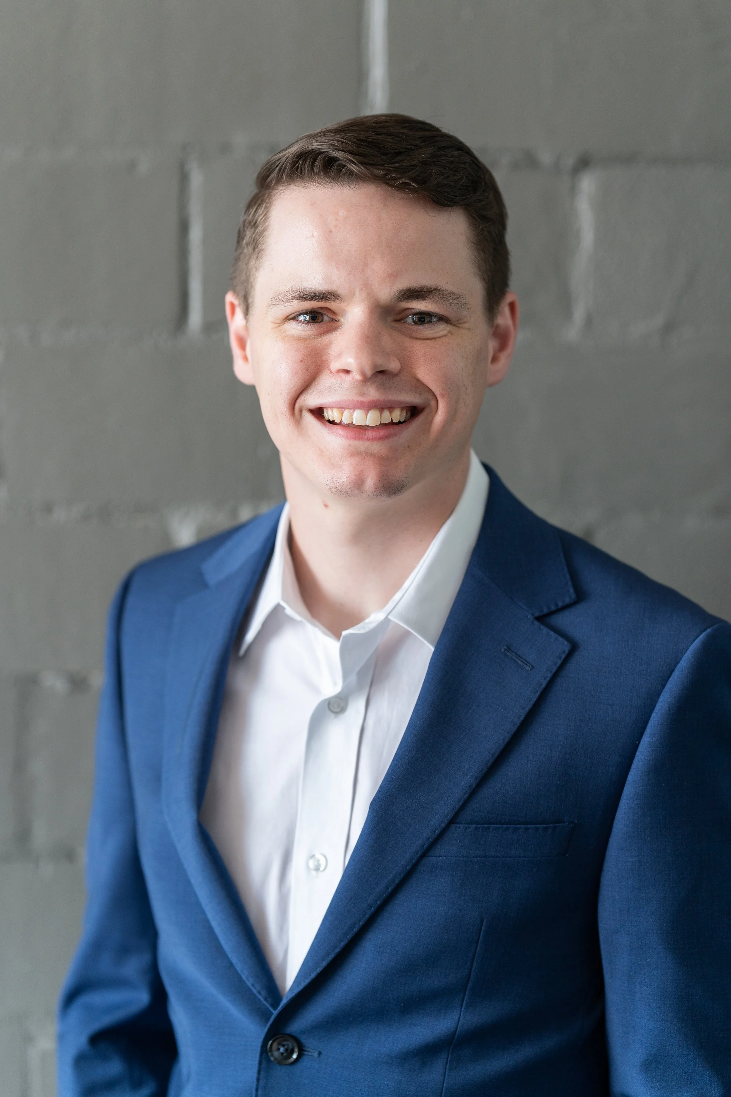 Portrait of a young man in a blue suit and white shirt, smiling against a gray concrete wall background.