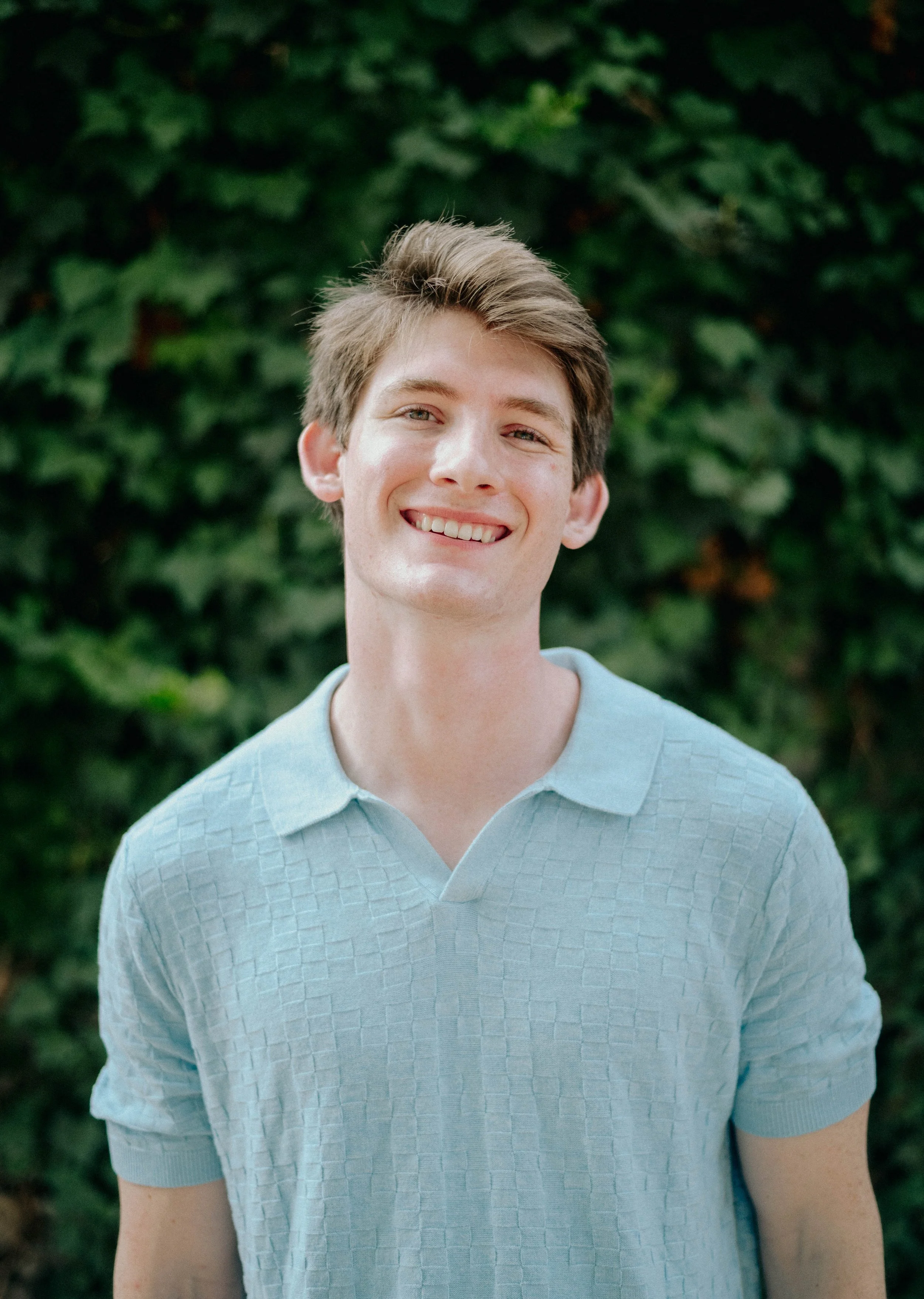 Young man with light brown hair smiling, wearing a light blue textured polo shirt, standing outdoors with green foliage in the background.