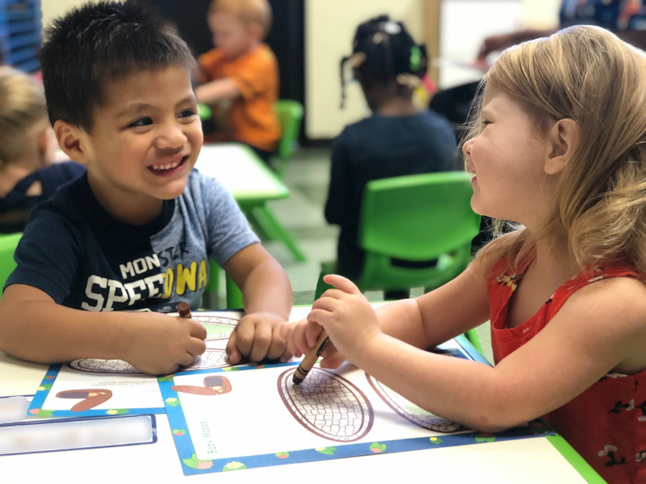 Two young children, a boy and a girl, sitting at a classroom table, smiling and engaging in a coloring activity with papers and markers. The background shows other children and classroom furniture.