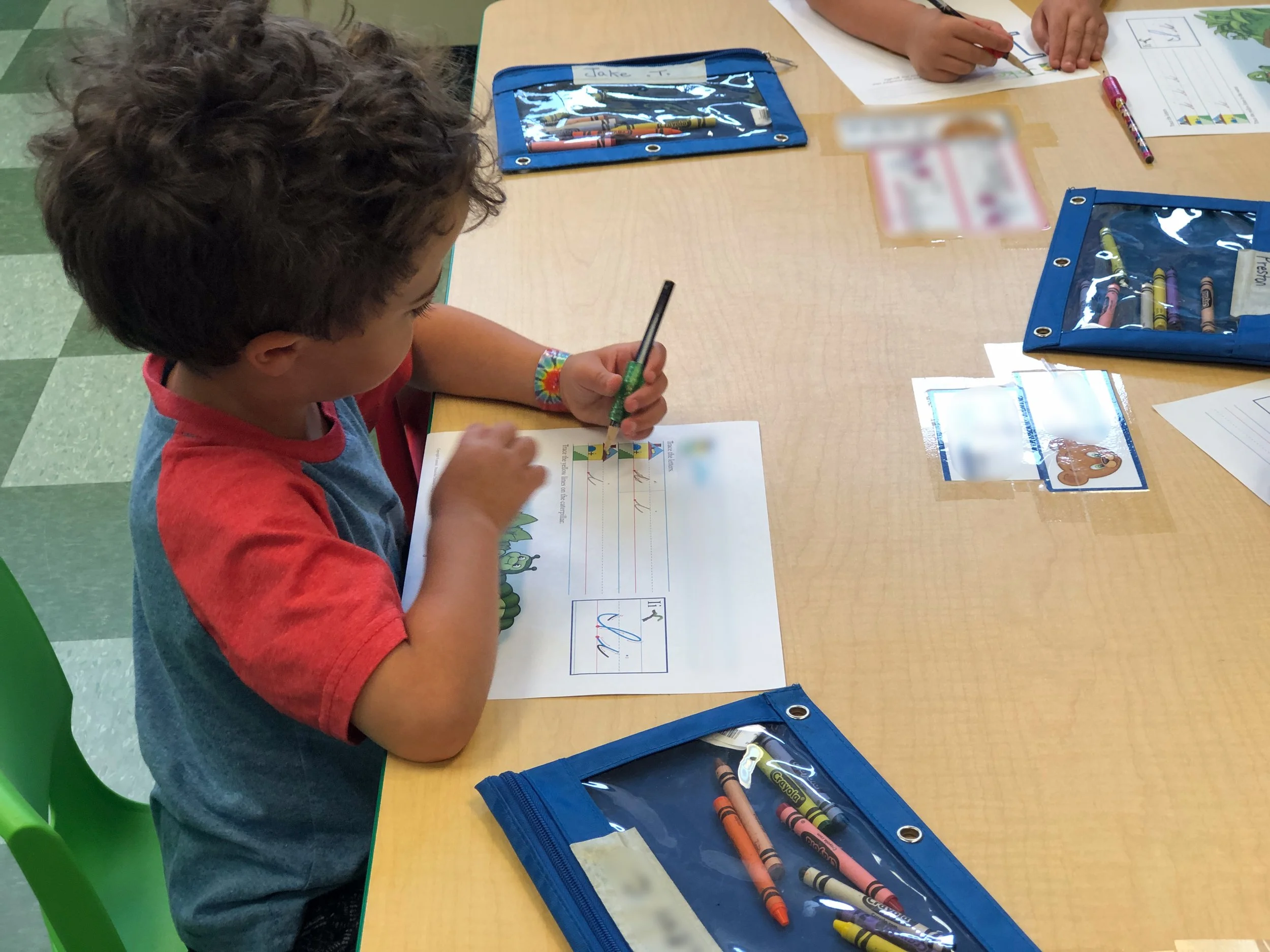 A young boy in a red and gray shirt sitting at a table, coloring a worksheet with crayons. There are blue crayon cases and other children's worksheets on the table.