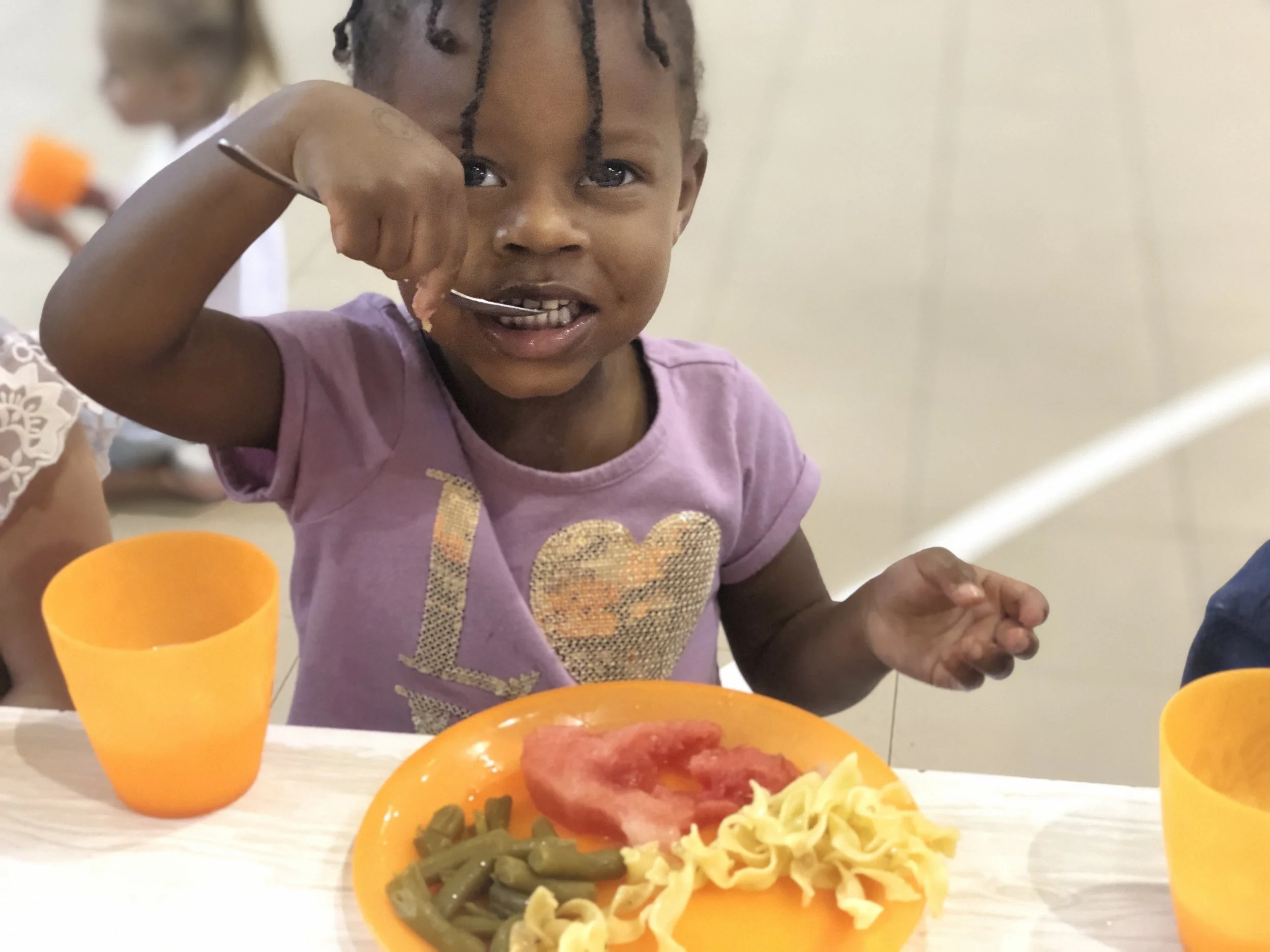 A young girl with braided hair, wearing a purple T-shirt with a glittery heart, is sitting at a table eating pasta, green beans, and a slice of tomato from an orange plate. She is smiling and has a fork in her right hand, with one piece of pasta on t