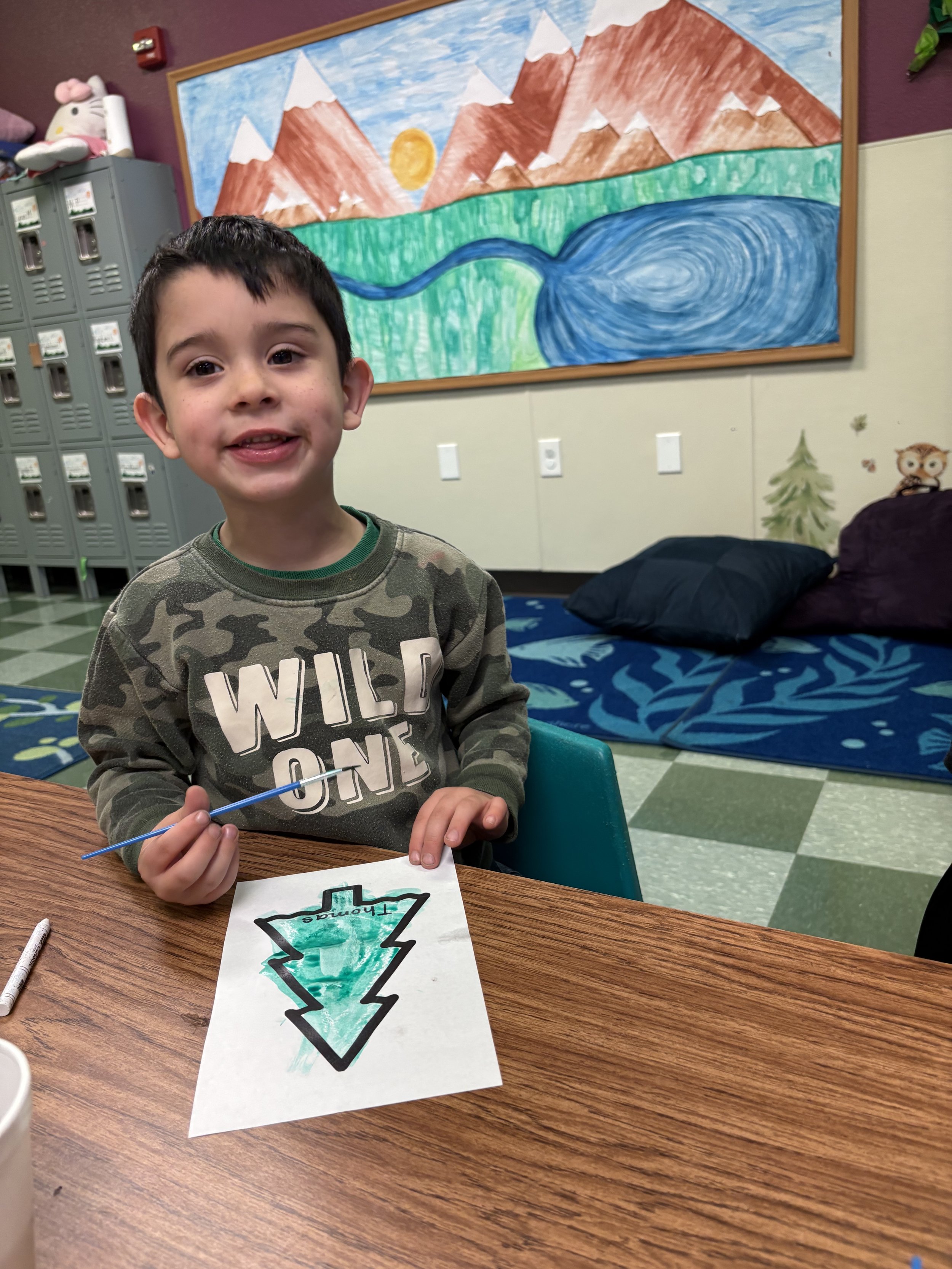 A young boy sitting at a wooden table in a classroom, smiling and holding a paintbrush with a drawing of a green Christmas tree on a piece of paper in front of him. Behind him is a mural of mountains, a lake, and a sun, along with lockers and plush t