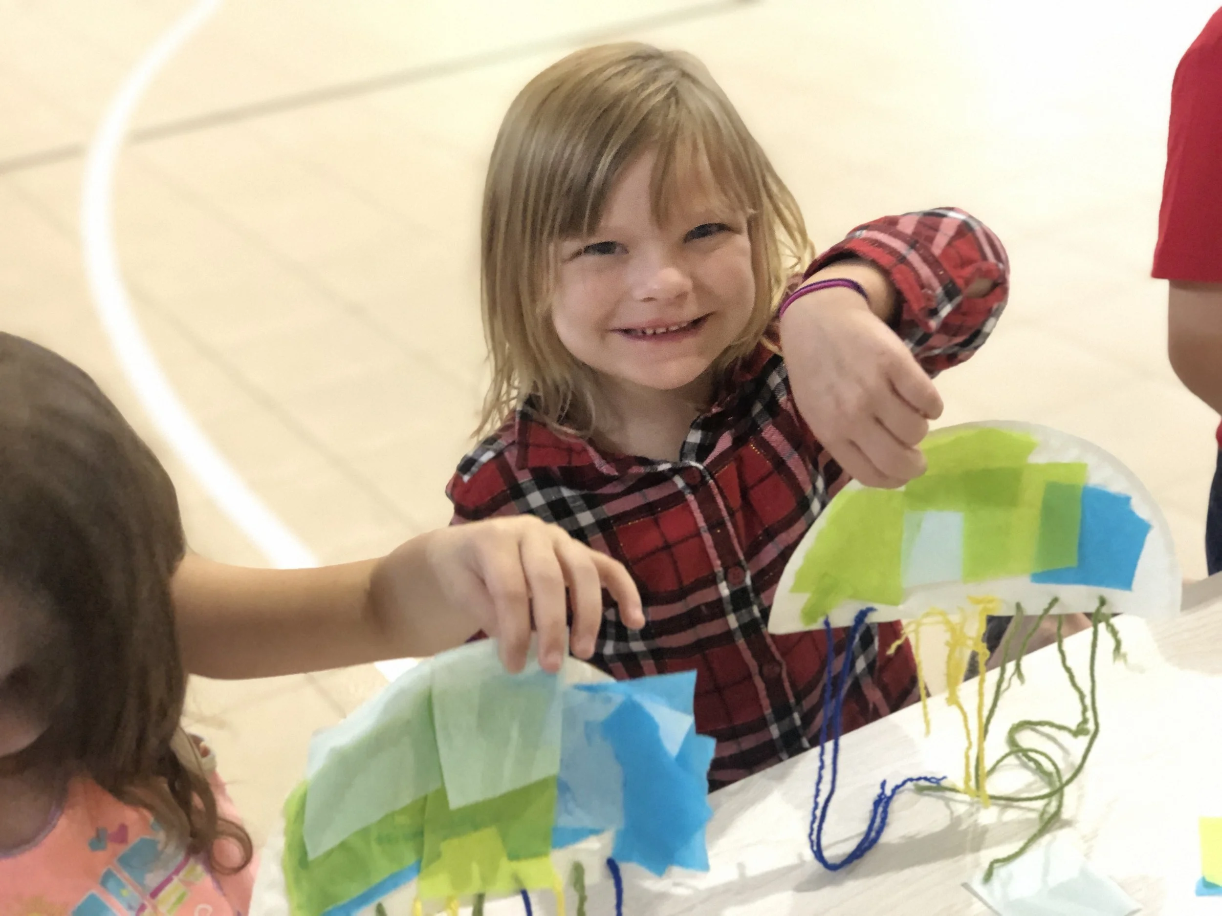 Smiling young girl with red hair wearing a plaid shirt, holding a colorful paper craft with green, blue, and yellow paper, and yarn strings.