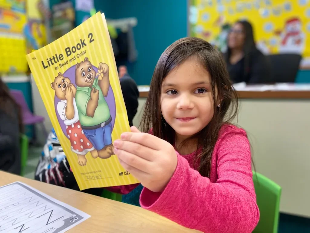 Young girl in a classroom holding a yellow workbook titled "Little Book 2 to Read and Color" with cartoon bear characters on the cover.
