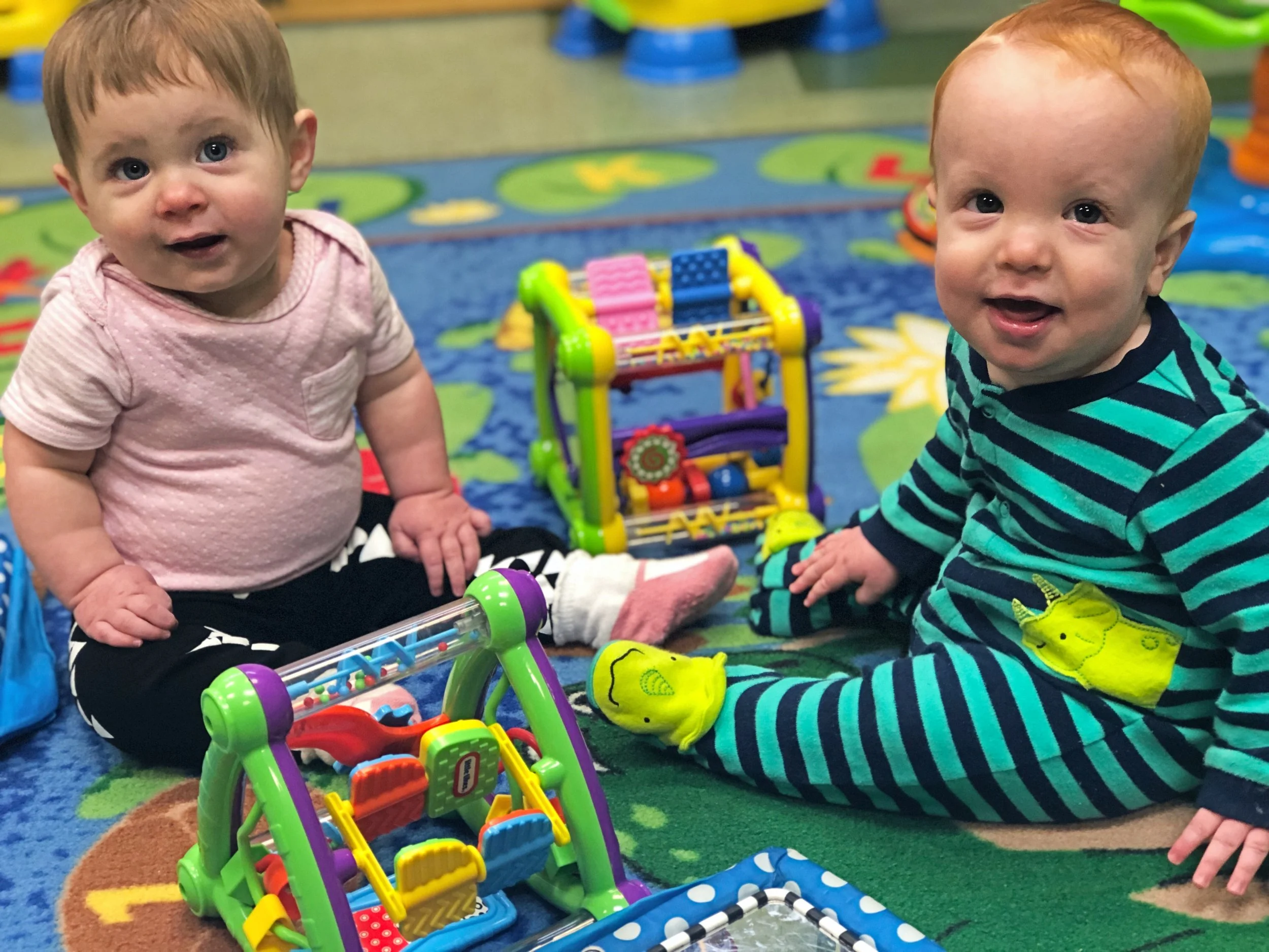 Two young children sitting on a colorful patterned play mat with toys, smiling at the camera in a playroom.