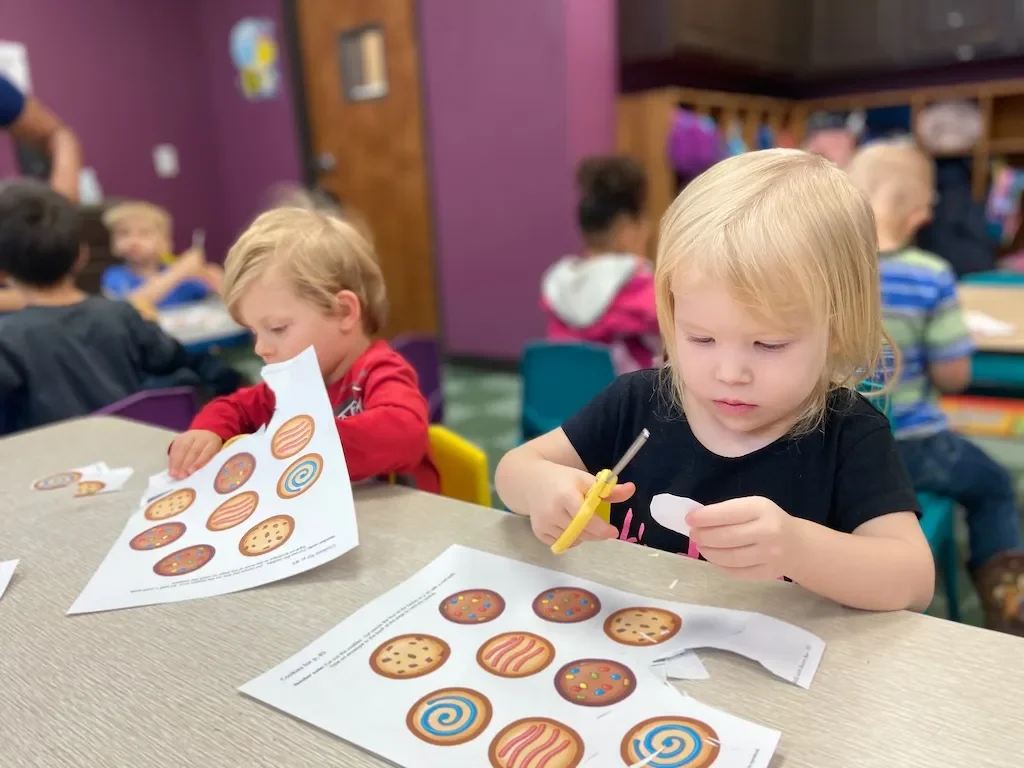 Two young children working on a cookie coloring activity in a classroom. The girl in a black shirt is cutting out a cookie shape, and the boy in a red shirt is looking at a paper with cookie illustrations.