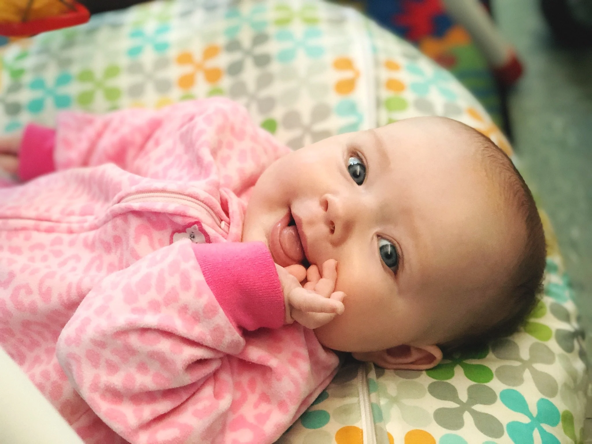 A baby lying on a colorful patterned blanket, wearing a pink leopard print hoodie, smiling with their tongue slightly out and hand near their mouth.