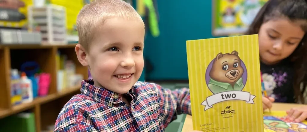Young boy smiling and holding a yellow educational book with a bear illustration, in a classroom with another child in the background.