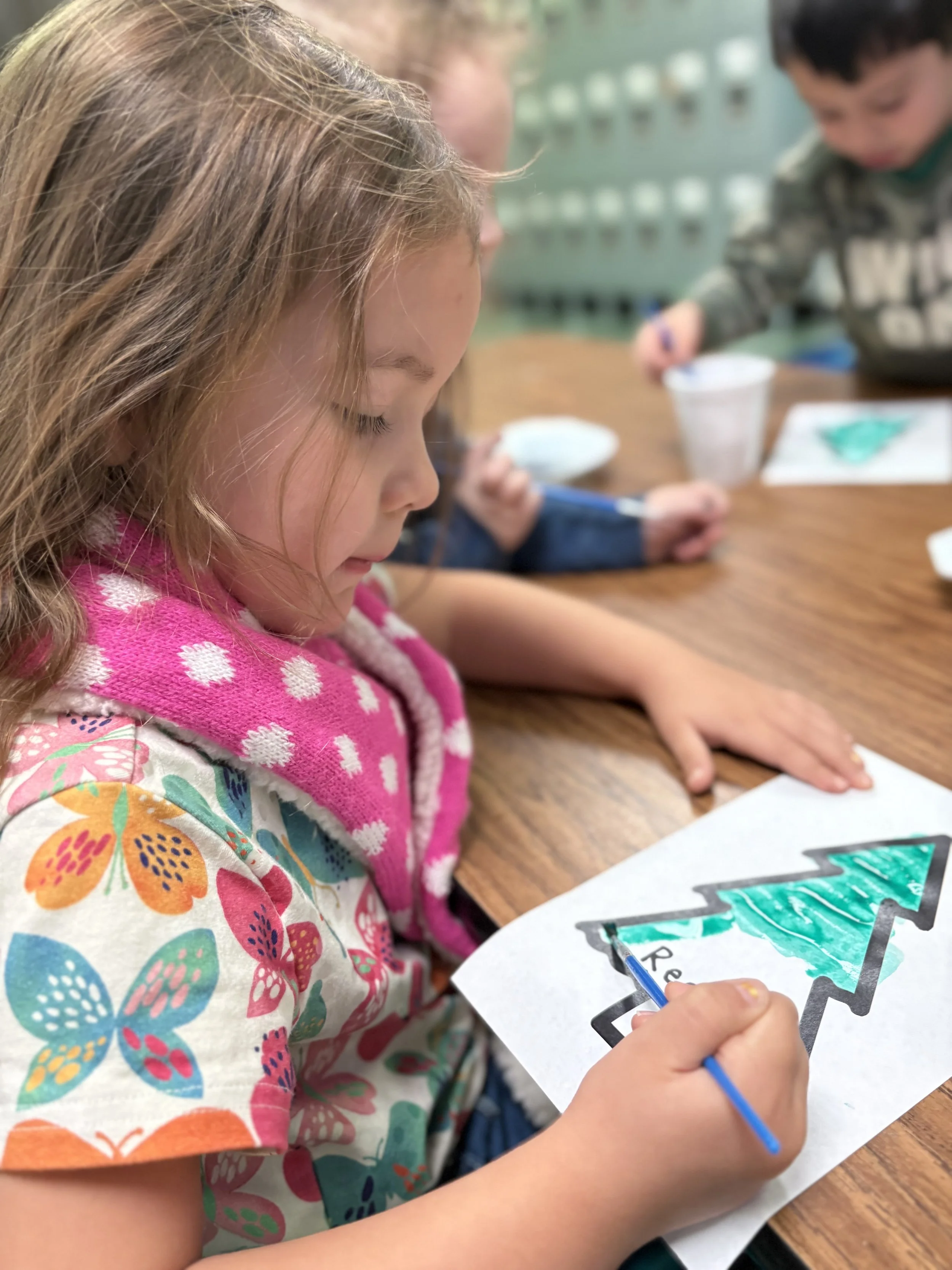 A young girl coloring a Christmas tree on a sheet of paper with markers, sitting at a wooden table with other children in the background.