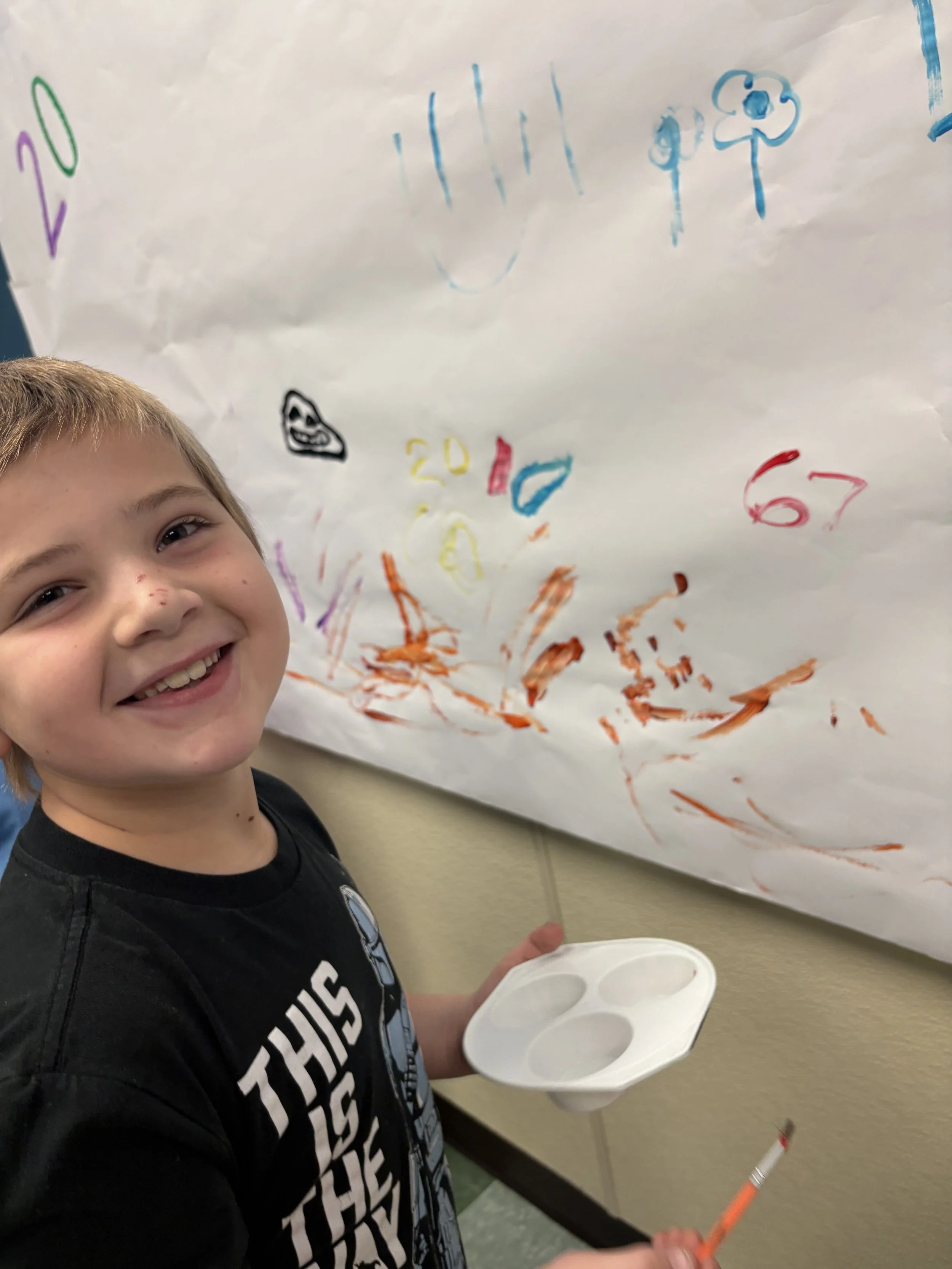 Young boy smiling and holding a paint palette with a paintbrush, standing next to a large sheet of paper with colorful drawings and scribbles.