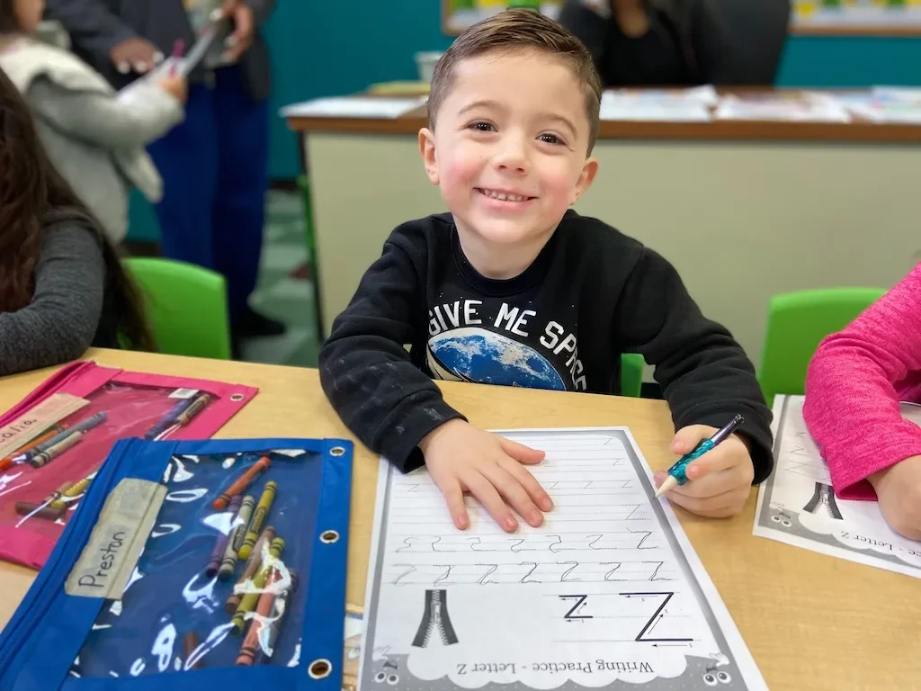 A young boy sitting at a desk, smiling, holding a pen, with lined paper and a worksheet with a large letter 'Z' on it, surrounded by colorful pencil cases, in a classroom.
