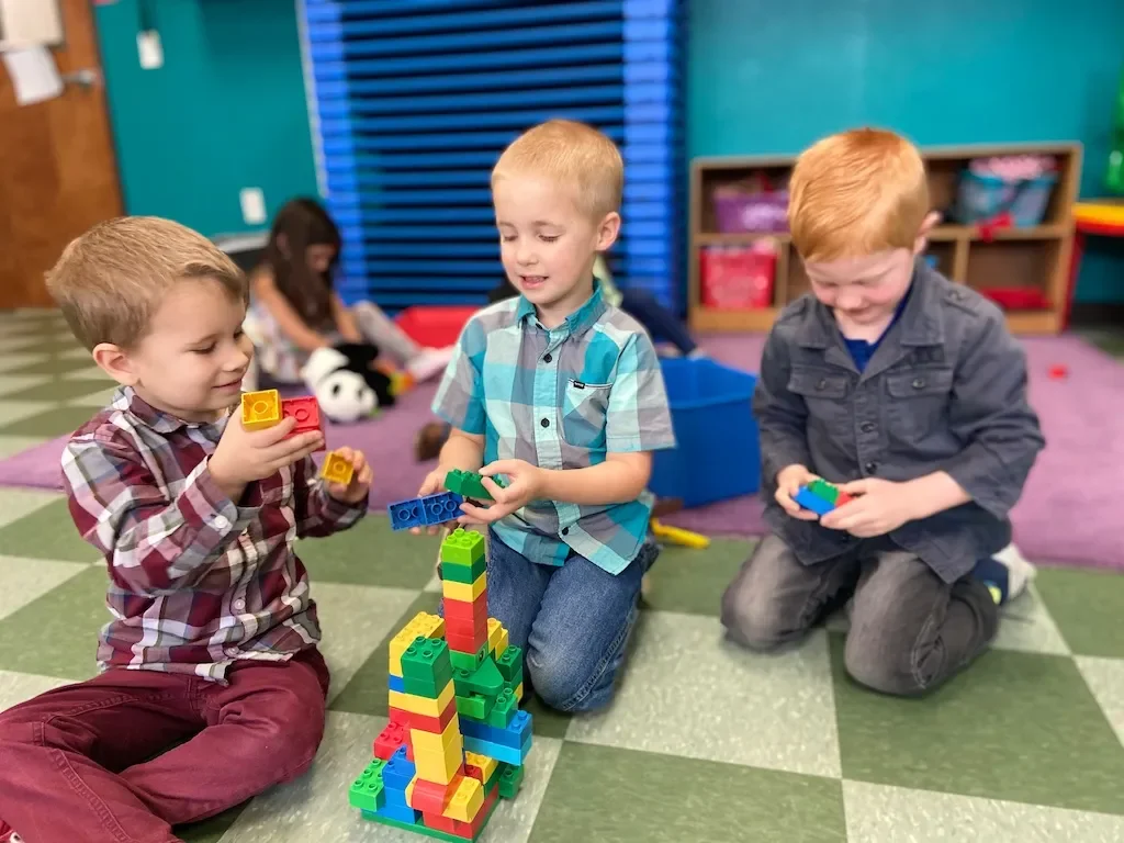 Three young boys playing with colorful interlocking building blocks on the floor in a classroom, with other children and shelves in the background.
