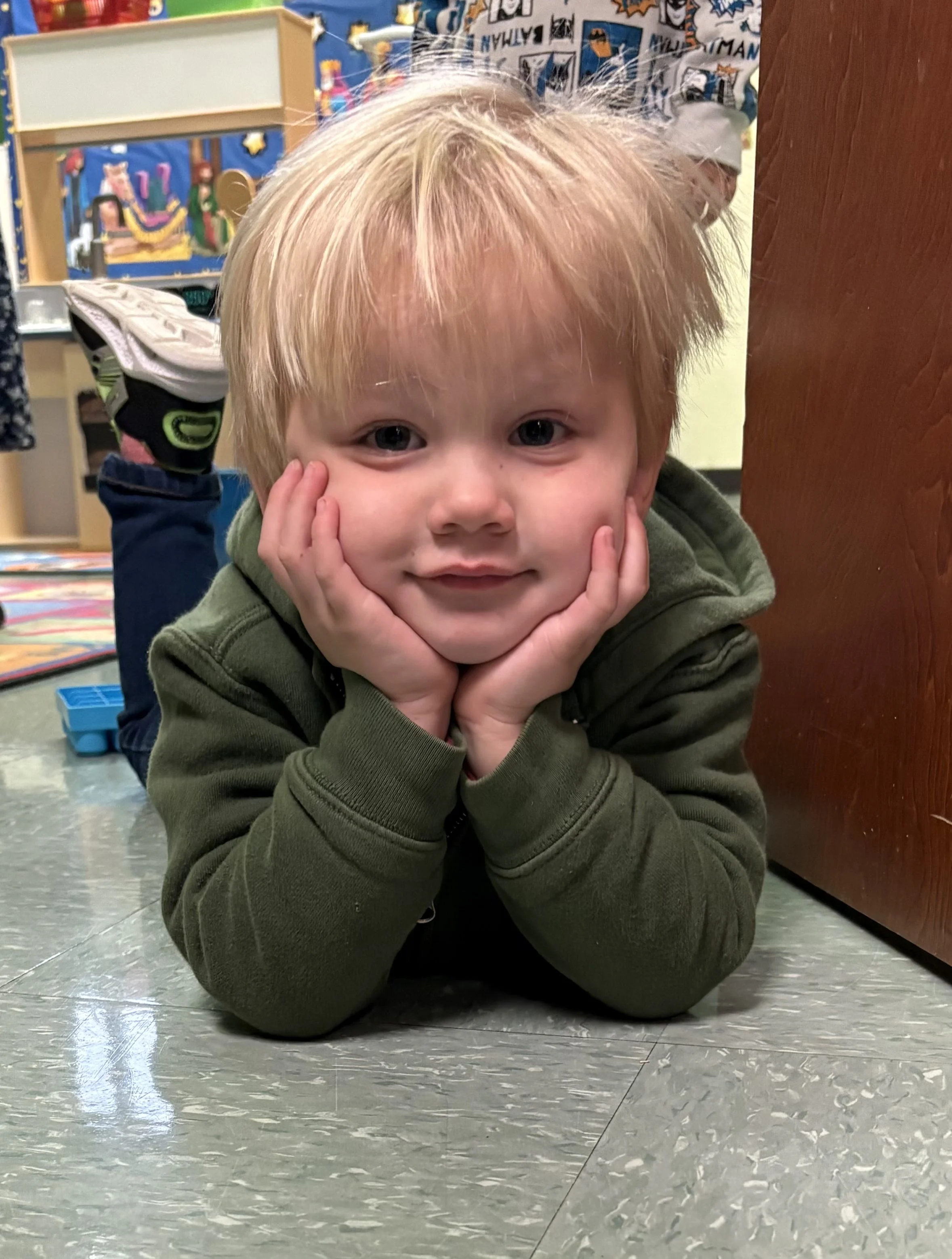 A young boy with blonde hair lying on his stomach on a checkered linoleum floor, resting his chin in his hands, smiling at the camera, in a colorful classroom.