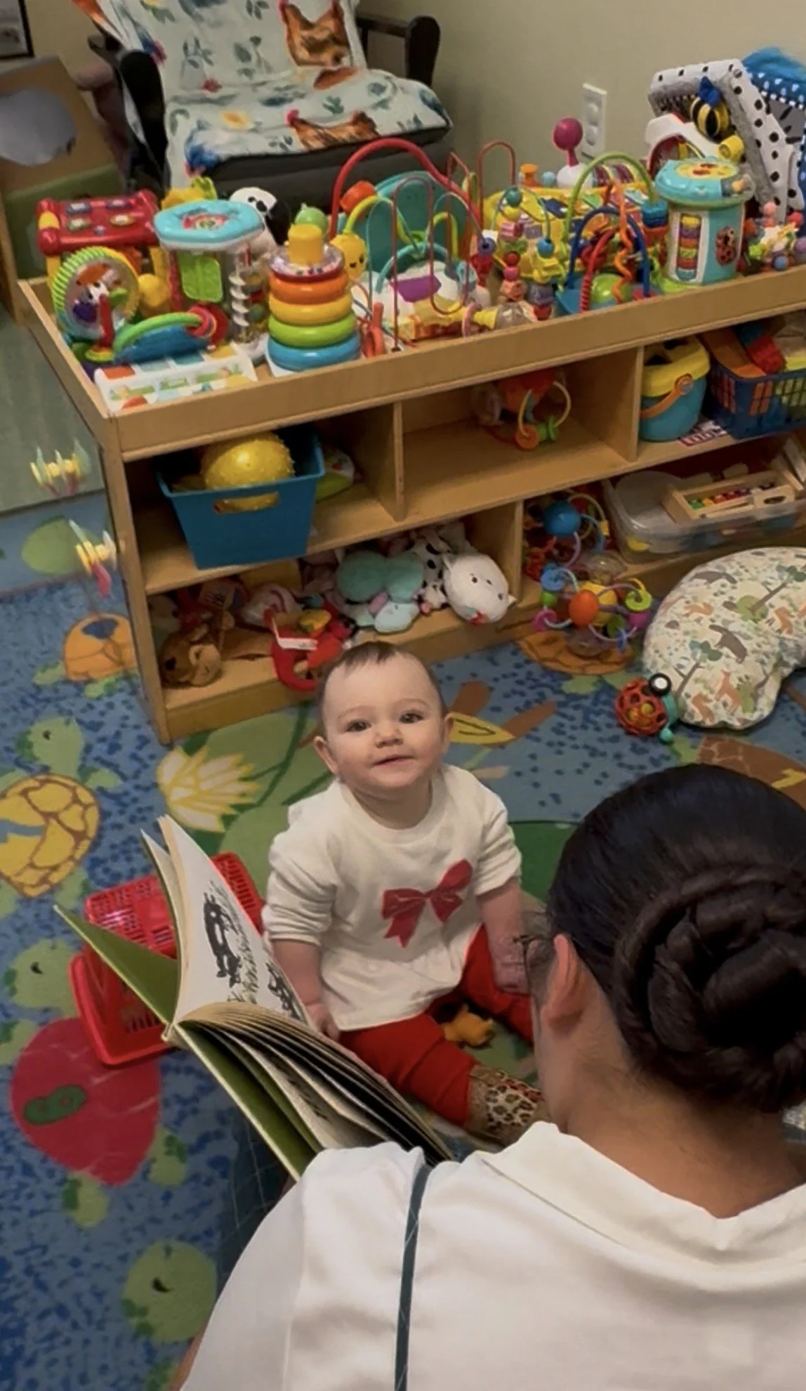 A young child sitting on a colorful animal-themed rug, looking up and smiling at an adult who is reading a book. In the background, there is a wooden shelf filled with various toys, including plush animals, activity toys, and stacking rings.