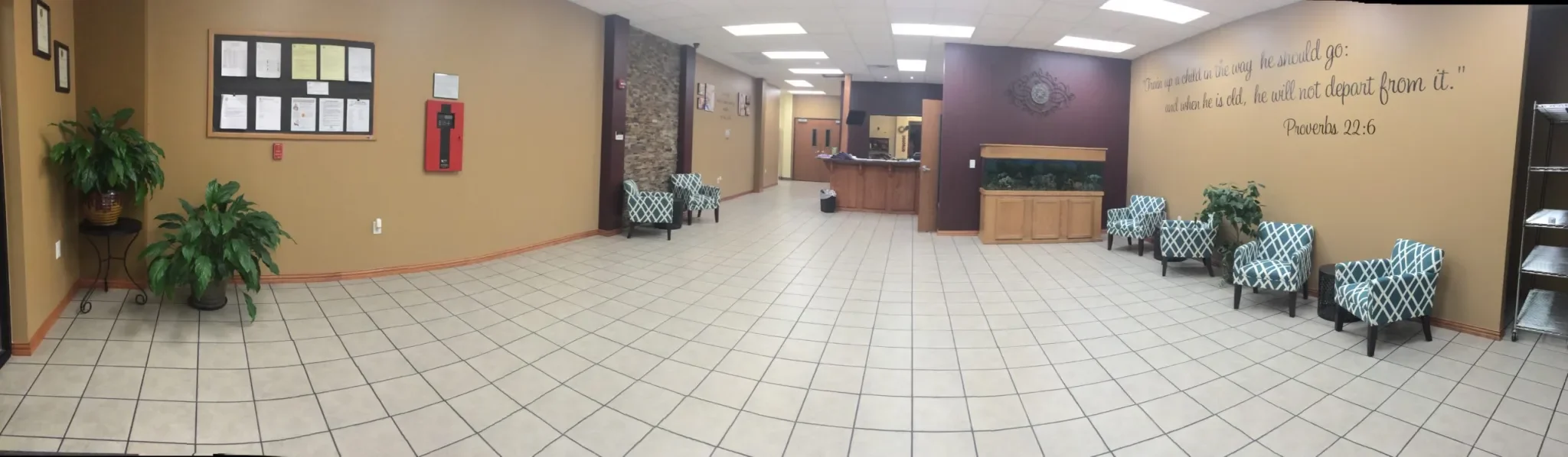 Spacious church lobby with patterned blue and white chairs, potted plants, framed notices on beige walls, and a quote from Proverbs 22:6 on the wall.