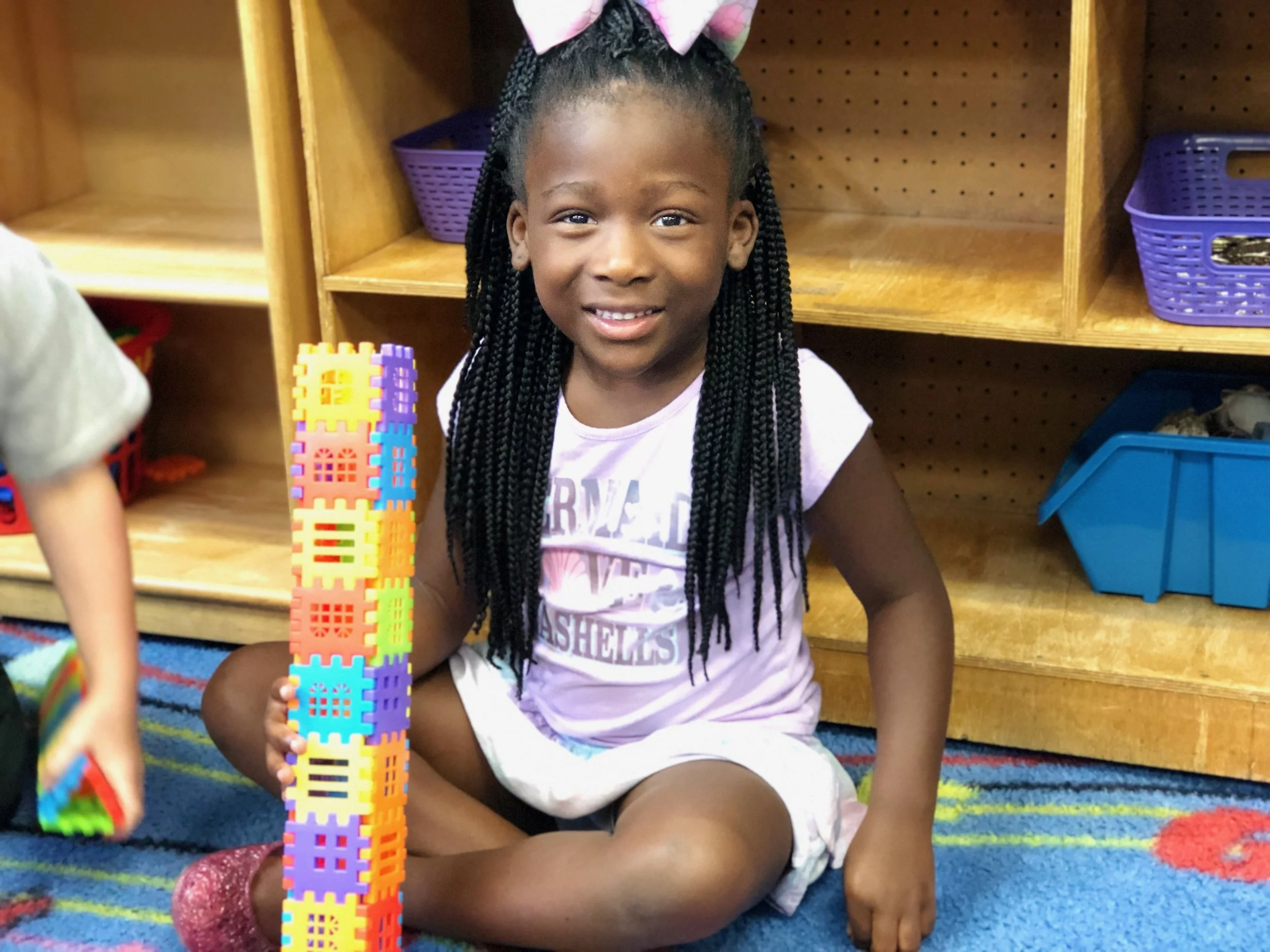 A young girl with braided hair, wearing a light pink shirt and shorts, sitting on a colorful carpet, playing with a tall, multicolored block tower, in a classroom setting with wooden shelves and storage bins in the background.