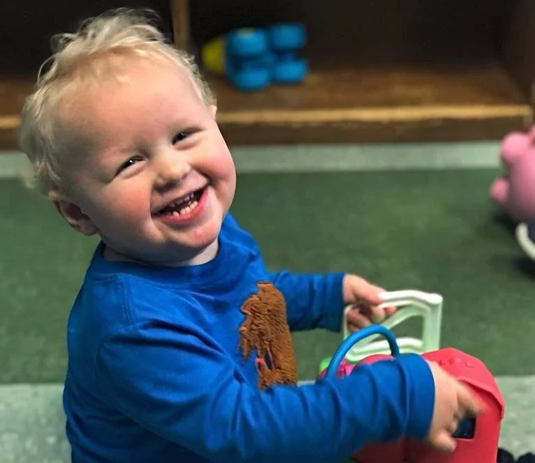 A young blond-haired boy in a blue shirt smiling while playing with a toy. There is a pink toy pig and some blue objects in the background.