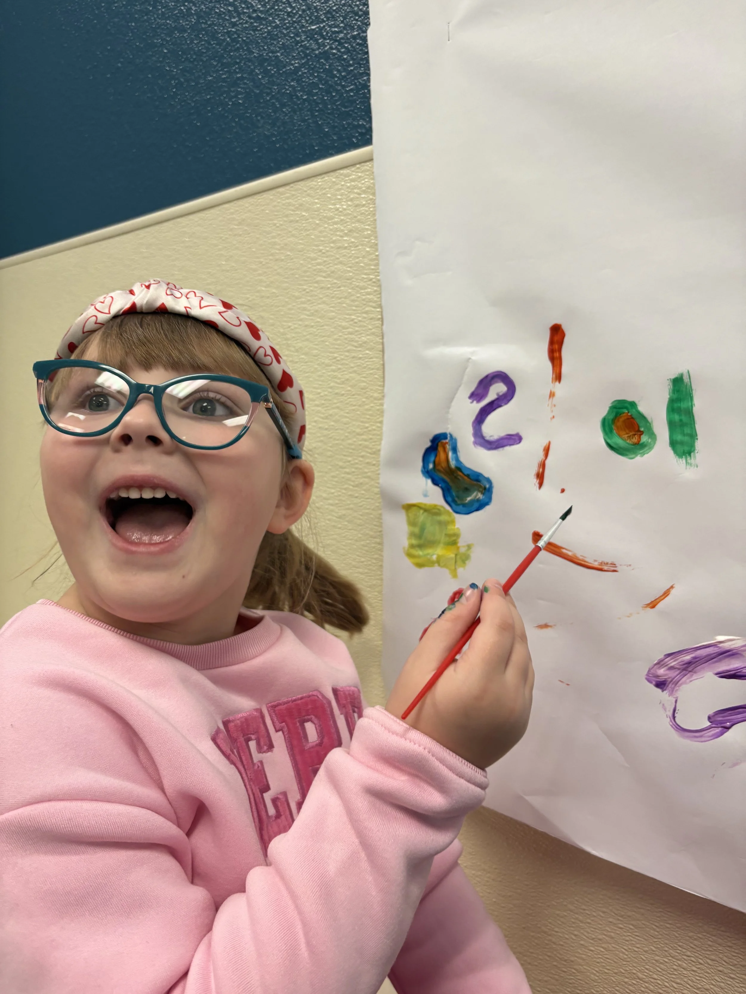 A happy girl with glasses, wearing a pink sweatshirt and a headband with hearts, painting on a large sheet of paper with bright colors.