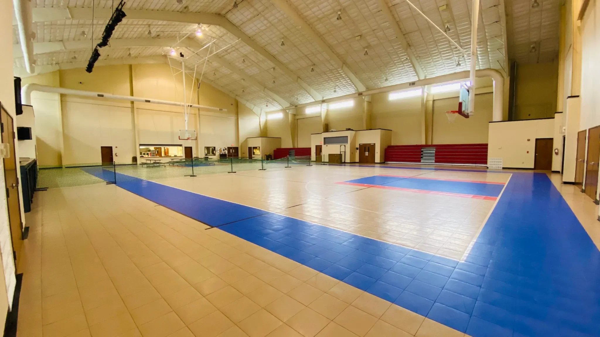Empty indoor gymnasium with a basketball court and a pickleball court, high ceiling, and red bleachers along the back wall.