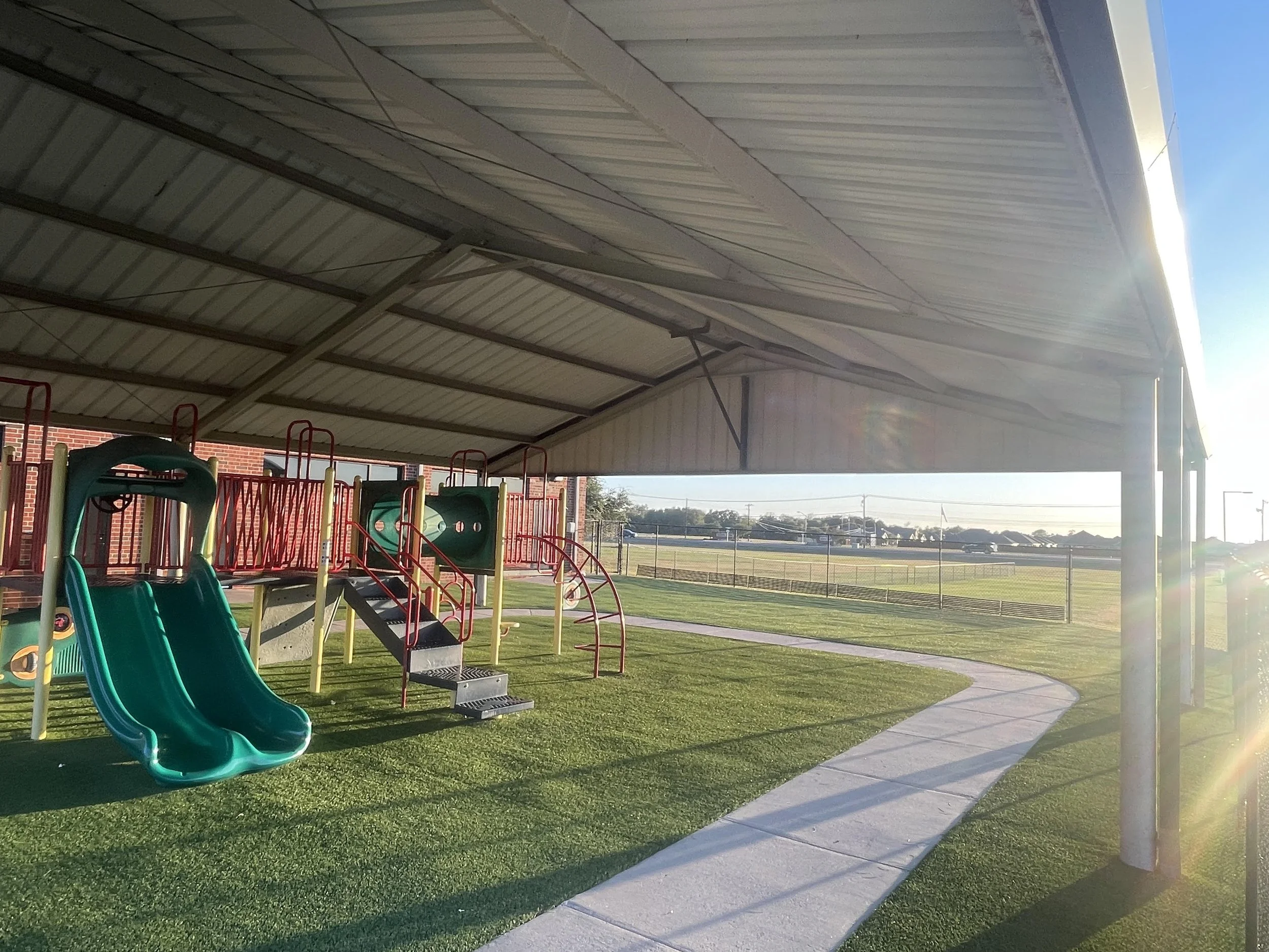 Colorful playground with slides and climbing equipment under a metal shelter, with a grassy field and fence in the background during sunset.