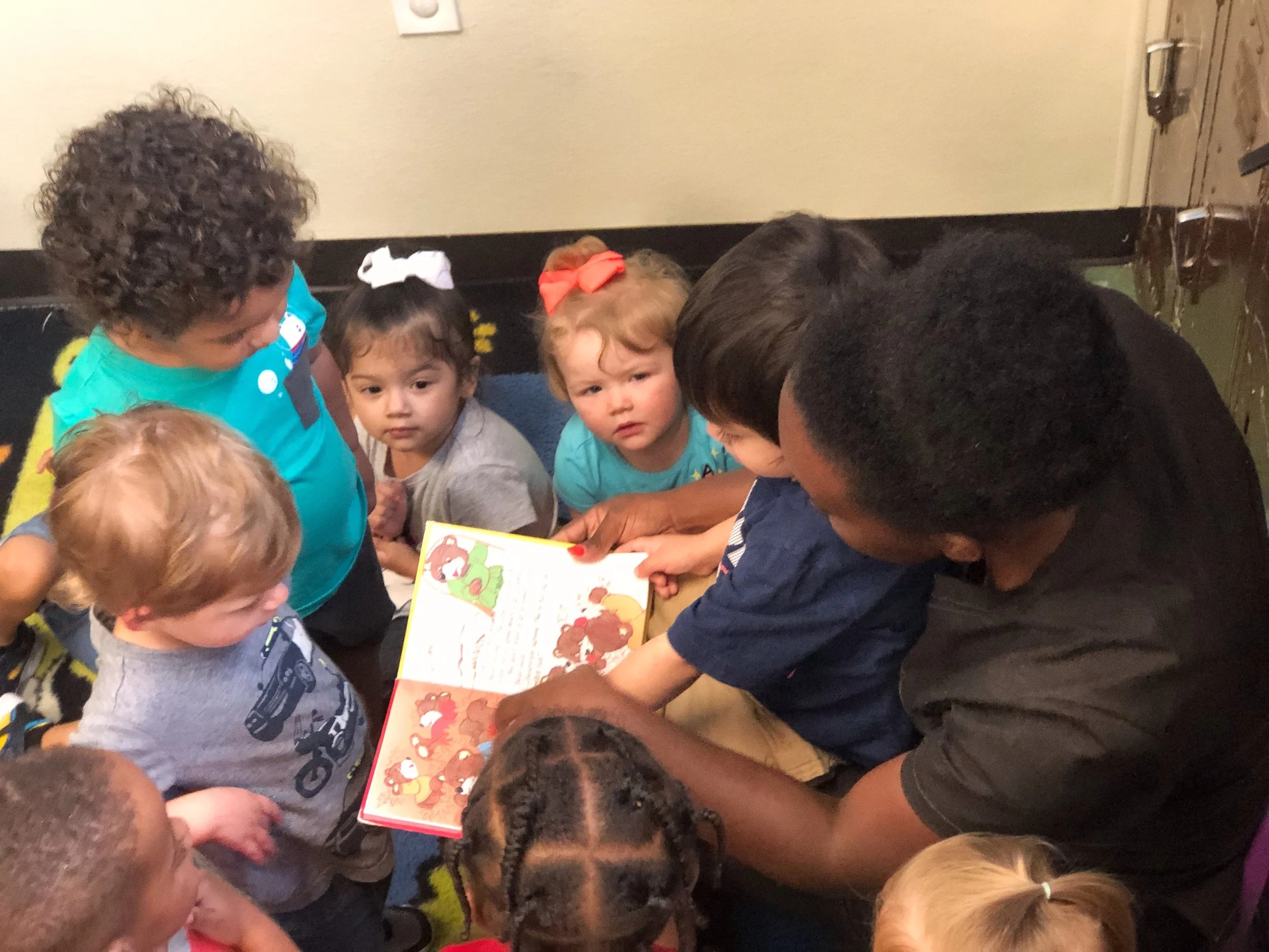 A group of young children with two adults sitting on the floor, looking at a colorful picture book together in a classroom or daycare setting.