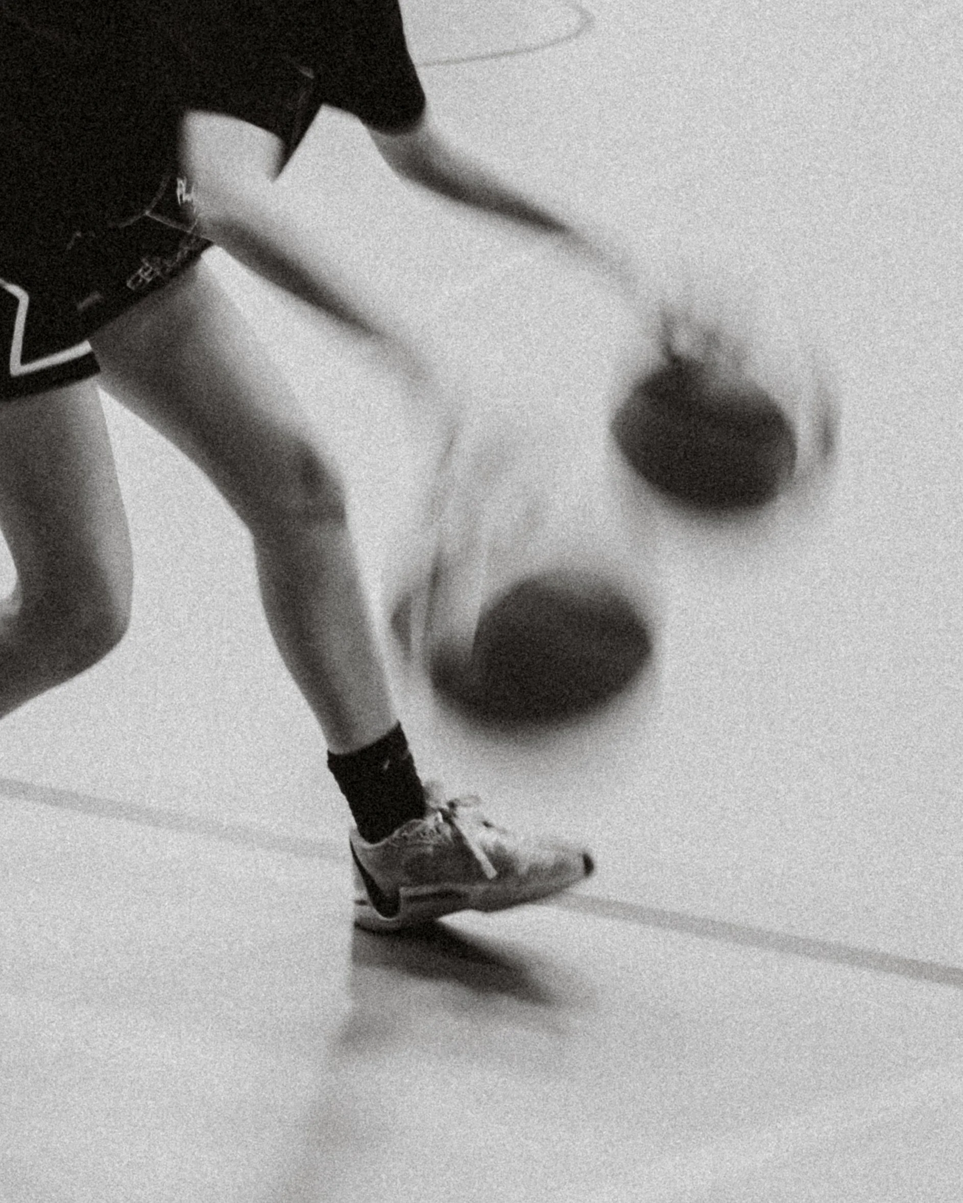 A person in athletic shorts and sneakers playing dodgeball indoors, with two dodgeballs in mid-air.