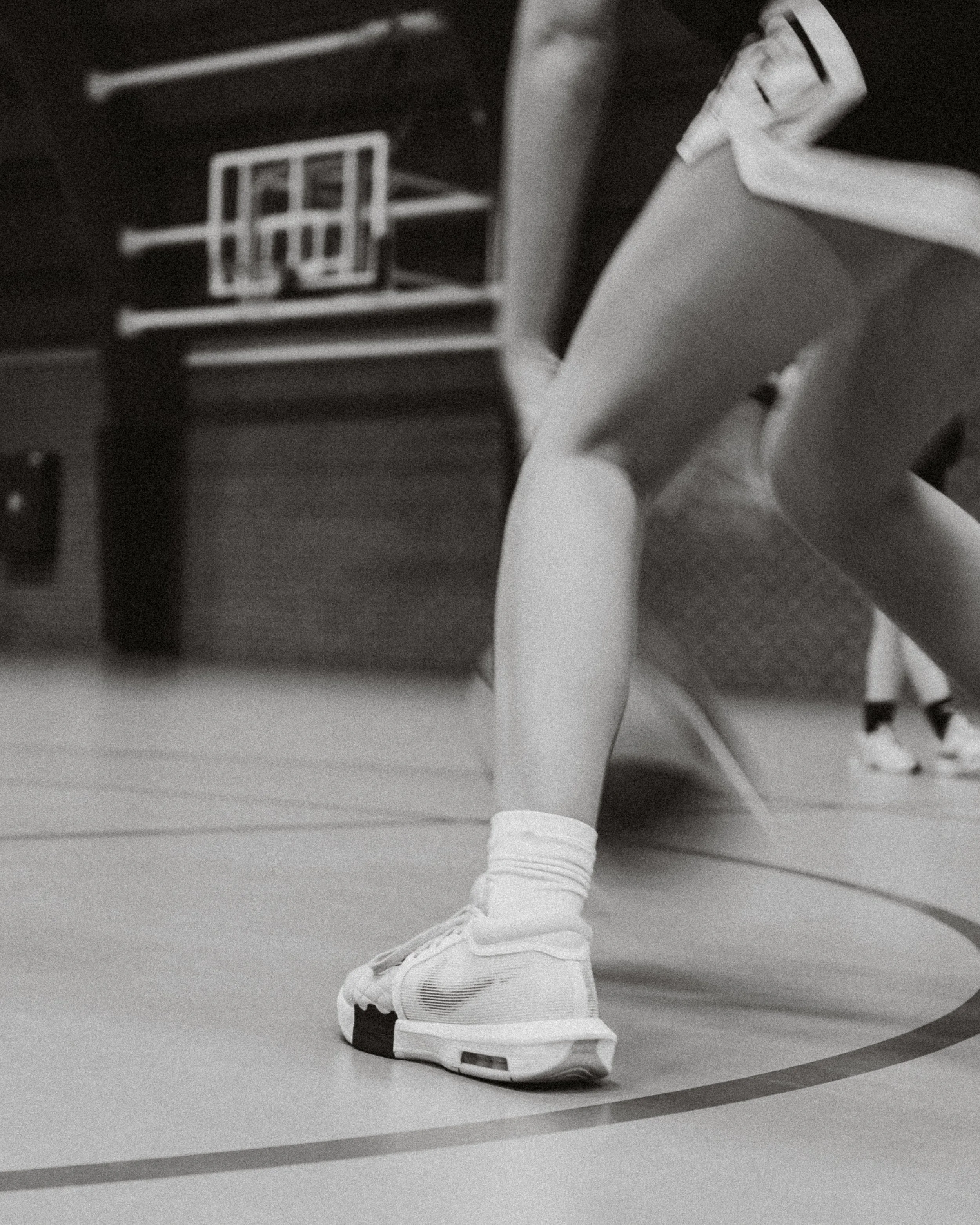 Close-up of a child's leg in a basketball uniform, wearing a sneaker, on a gym floor with a basketball hoop and backboard visible in the background.