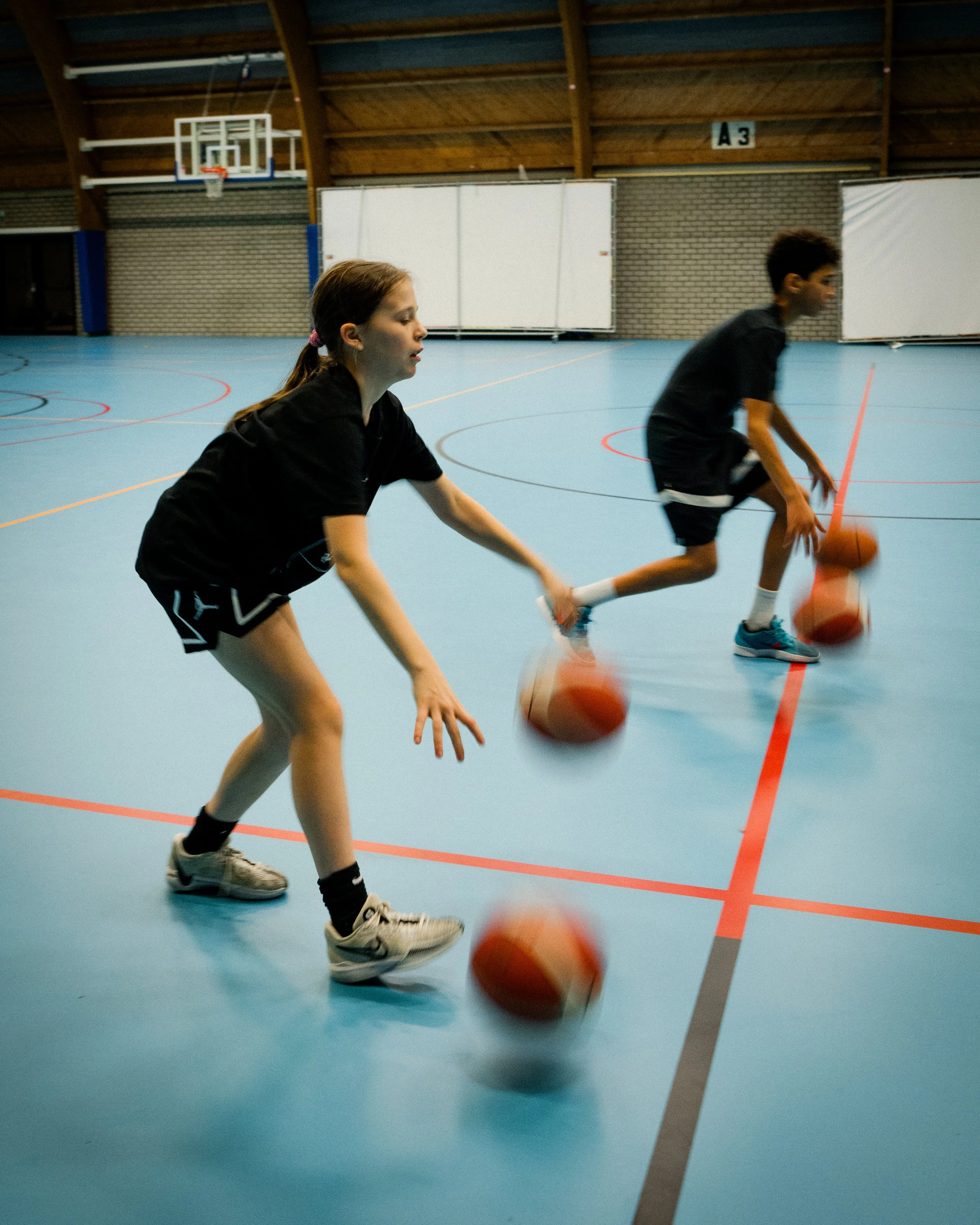 Two children practicing basketball drills in an indoor gym.