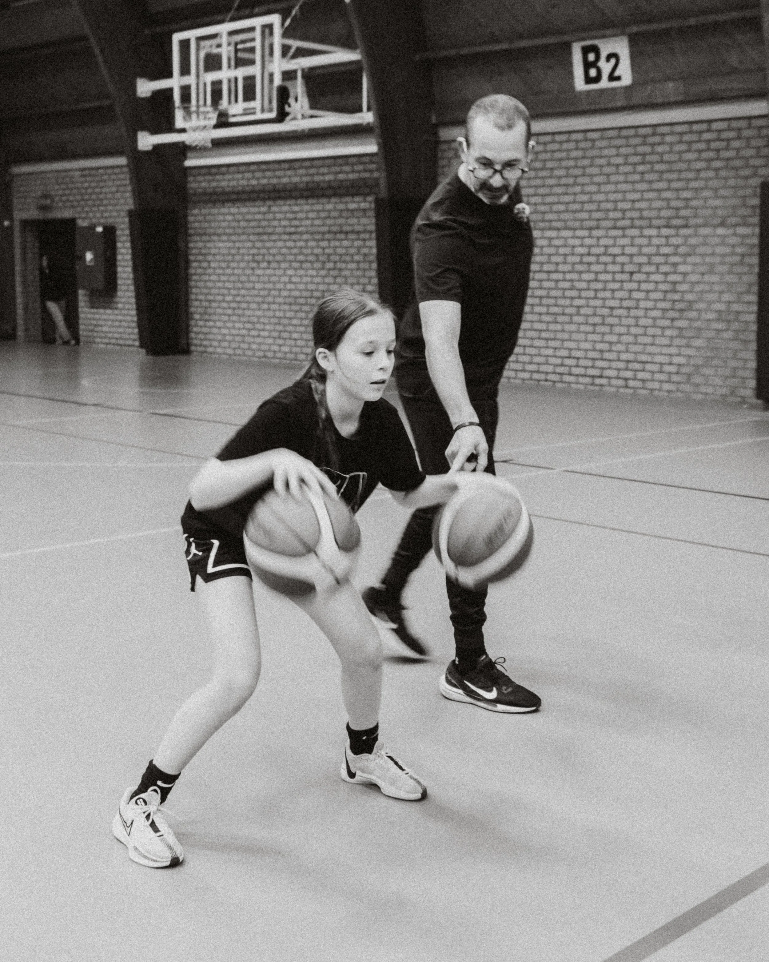 A young girl dribbling two basketballs in an indoor gym under the supervision of a coach.