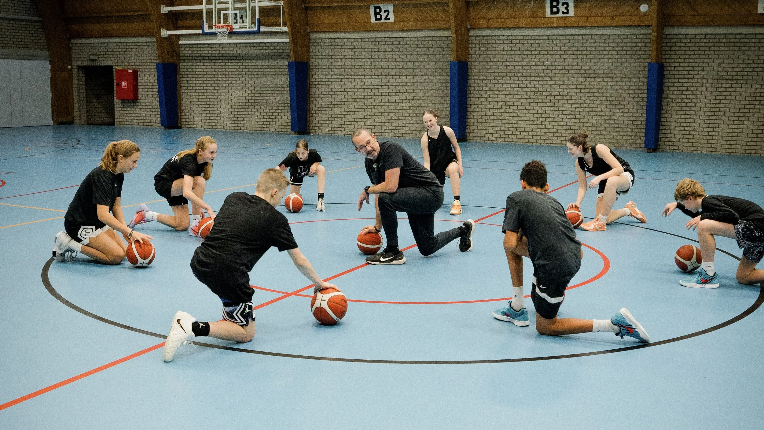 A basketball coach instructs young basketball players during a practice session in Ter Groene Poorte, Brugge. The players are kneeling on the blue court, each with a basketball, and are listening attentively to the coach.