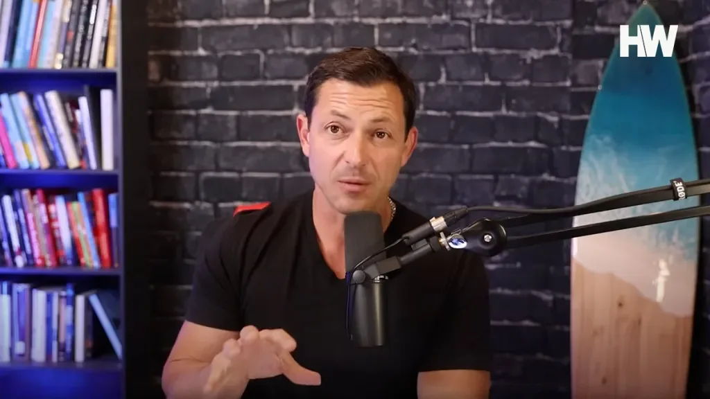 A man speaking into a microphone in front of a brick wall with a bookshelf on the left and a surfboard on the right side.