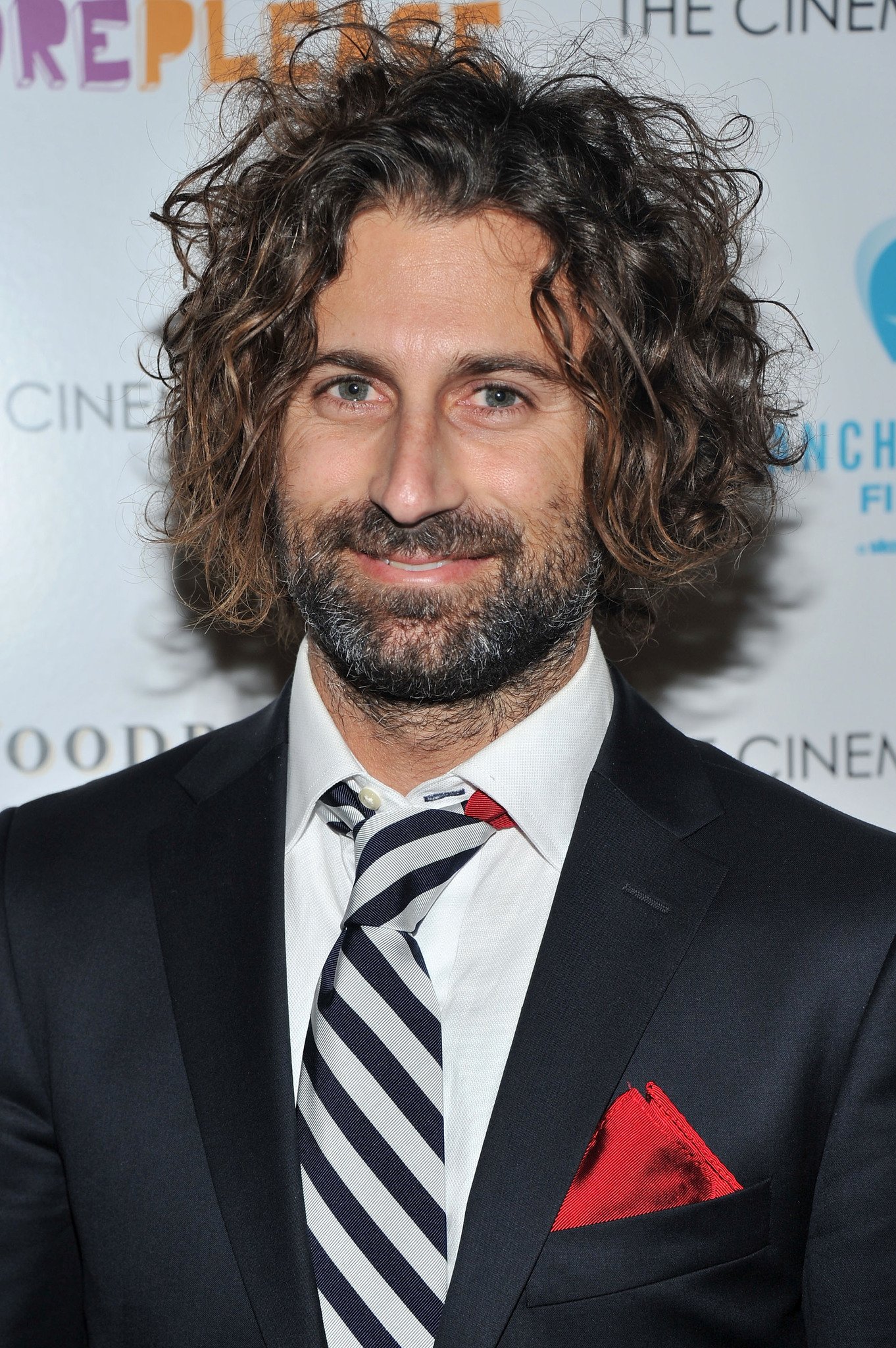 A man with curly brown hair and a beard is smiling at the camera, dressed in a black suit with a white shirt, a striped tie, and a red pocket square. The background includes a partially visible event or film festival backdrop.