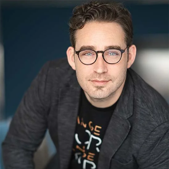 A man with dark, styled hair, glasses, and a light beard, wearing a dark blazer and graphic t-shirt, sitting indoors against a blue background.