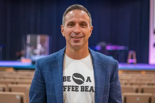 A smiling man in a blue blazer and a white t-shirt with the text 'BE A COFFEE BEAN' stands in an auditorium with empty seats and a stage in the background.